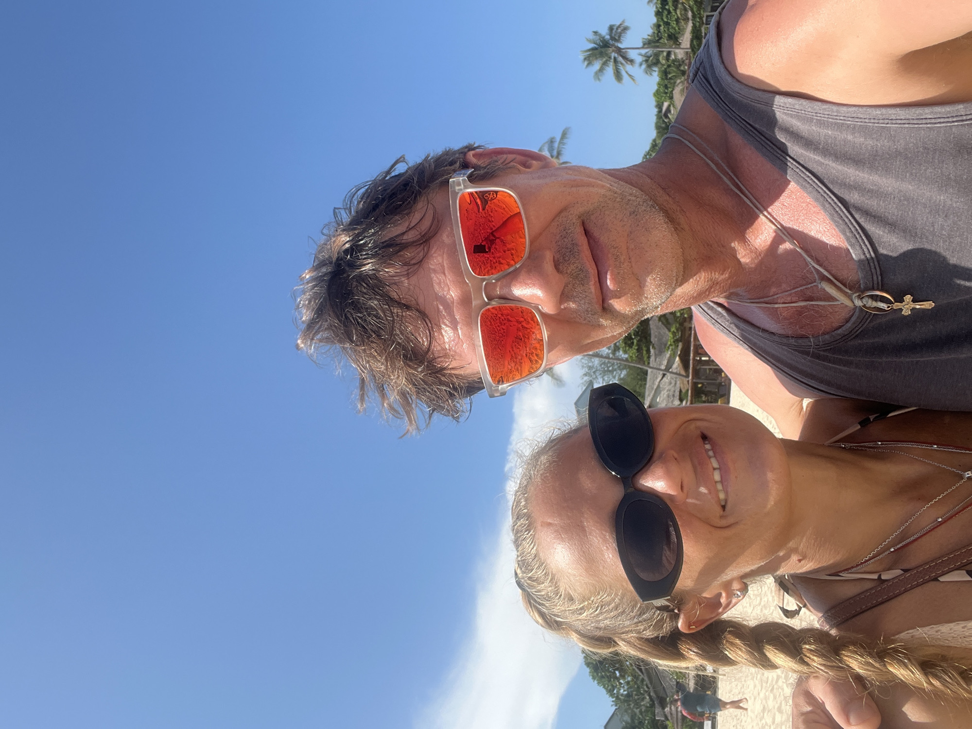 This is a selfie taken on a beach with two people, a man and a woman, in the foreground. Both are wearing sunglasses and smiling. The man has messy hair and is wearing a grey tank top with a necklace. The woman has blonde hair in a braid and is wearing a patterned top and necklaces. Behind them, there are palm trees, some buildings, and a clear blue sky with a few clouds. The man's sunglasses reflect the camera and a circular emblem. The background suggests a tropical location. The image does not depict the city of Phu Thuy Ward, Vietnam.