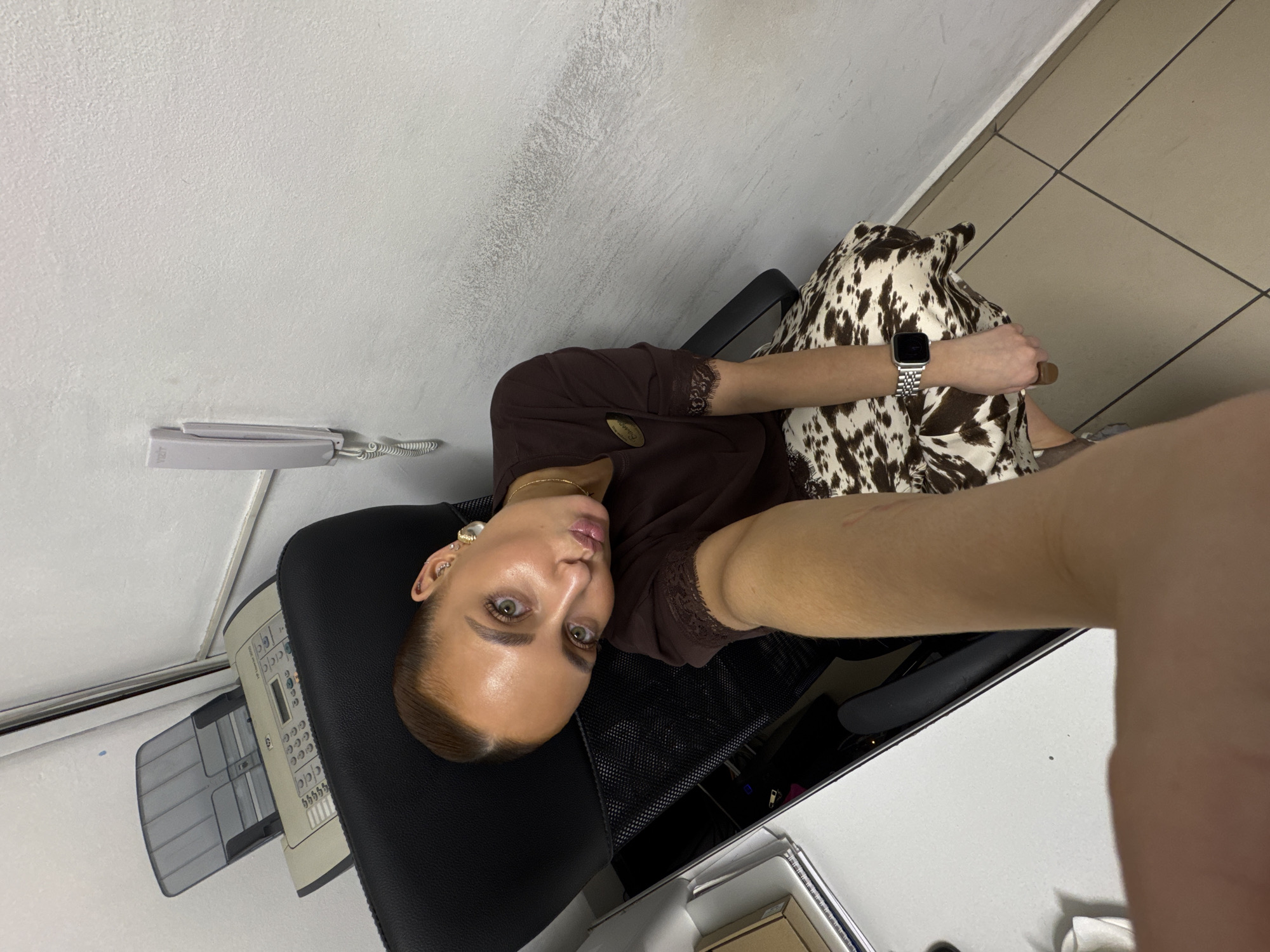 A woman is sitting in an office chair in what appears to be an indoor office environment. She is wearing a brown shirt with lace detailing and a silver wristwatch. She is holding her arm out towards the camera as if taking a selfie. Behind her is a pillow or cushion with a brown and white cow print. The background shows a wall with an intercom system, a desk with office supplies, and tiled flooring. While the image hints at an office setting, there is no verifiable evidence within the image to confirm that the location is indeed the city of Chaikovsky, Russia.