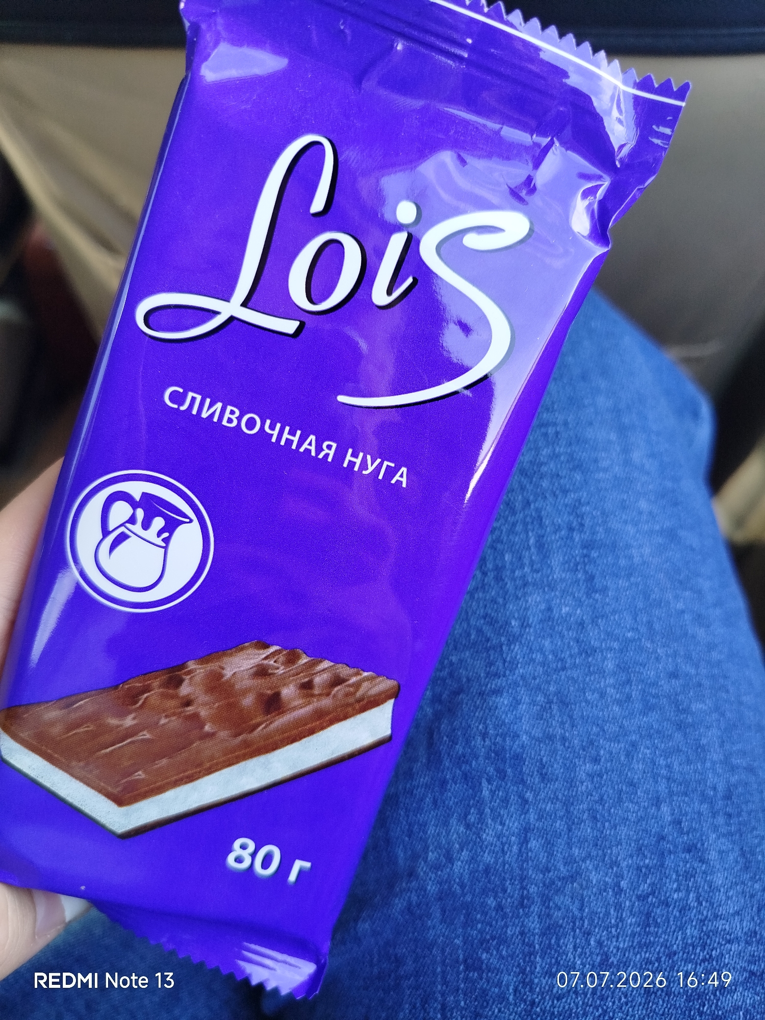 A close-up shot captures a purple chocolate bar wrapper with the white text "Lois" prominently displayed in a script font. Below the brand name, in smaller white text, is "СЛИВОЧНАЯ НУГА," which translates to "creamy nougat." To the left of this text, there's a circular white logo depicting a pitcher with milk pouring out. The bottom of the wrapper shows a graphic of the chocolate bar itself, revealing its nougat filling. The weight "80 г" is printed in white text on the lower right of the wrapper. The image also includes text overlays indicating "REDMI Note 13" and the date "07.07.2026 16:49." The chocolate bar is held by a person, and the background shows parts of blue denim clothing. The scene appears to be indoors, possibly in a car, given the muted, blurred background. There is no visible indication of the city of Nizhny Bestyakh, Russia.