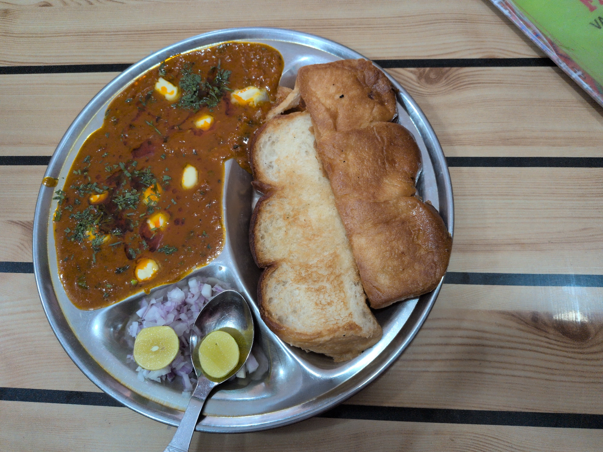 The image shows a stainless steel thali plate containing a portion of Pav Bhaji, a popular Indian street food. The Pav Bhaji consists of a thick vegetable curry topped with dollops of butter and chopped cilantro, served in one section of the thali. Adjacent to the curry are two pieces of toasted pav (bread rolls), golden brown and slightly puffed. In another section of the thali, finely chopped red onions are visible, garnished with two thin slices of lemon. A spoon rests in the onions, also holding a lemon slice. The thali is placed on a wooden table with visible grain patterns and black vertical lines. The lighting appears to be overhead, casting a soft glow on the food and table. There are no people visible in the image. The setting suggests an eatery, likely a street-side stall or a casual restaurant in Vashisht, India.