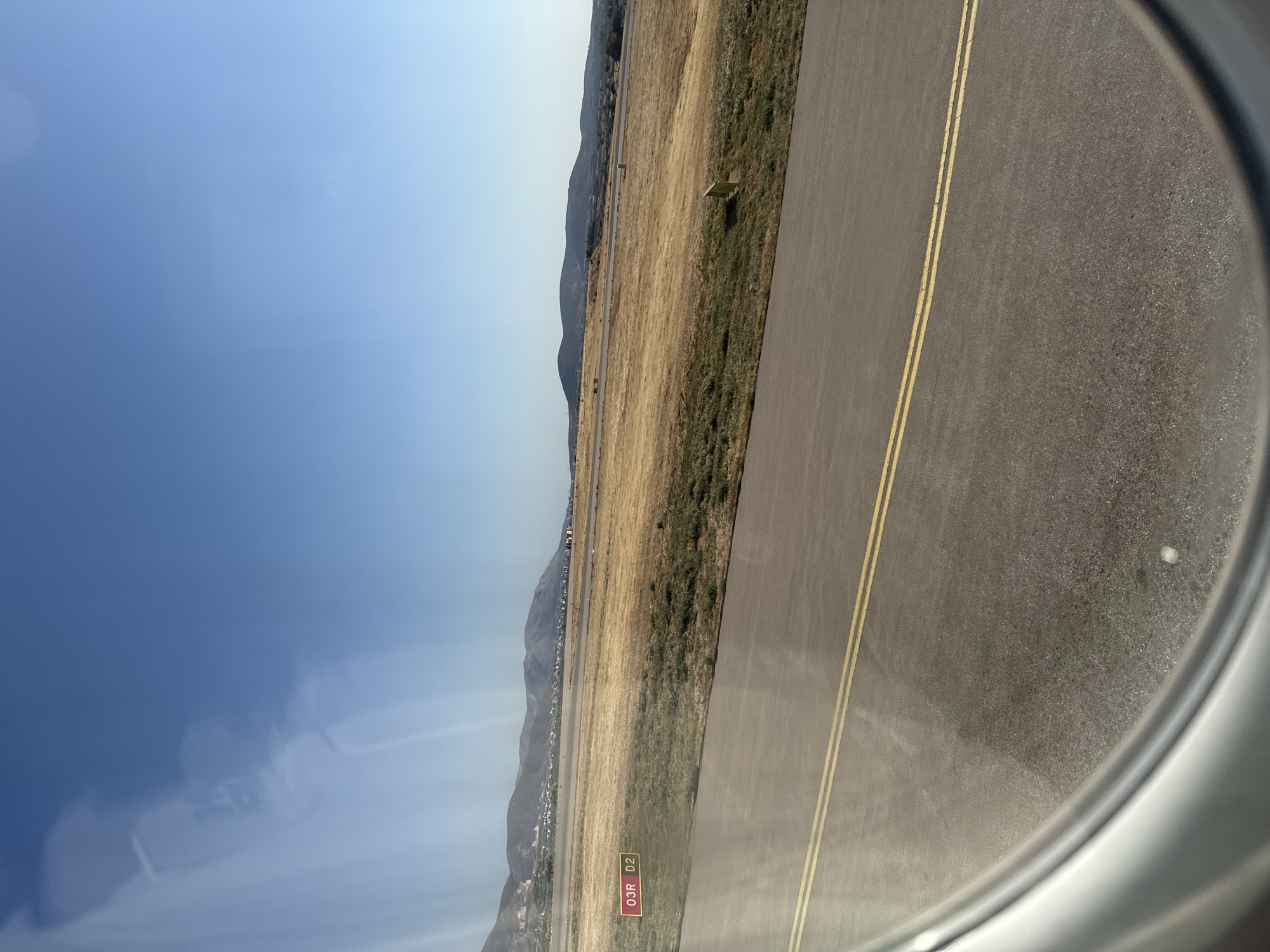 This image depicts an airport runway seen through an airplane window. The foreground is dominated by the asphalt runway with two yellow lines running parallel. Beyond the runway, there's a dry, grassy field stretching towards a line of low hills in the distance. The sky is clear and blue. A red and yellow sign with the text "03R D2" is visible on the left side of the runway. There are no people or vehicles visible in the frame, suggesting the scene is either before takeoff, after landing, or during a quiet period at the airport. The environment appears to be arid or undergoing a dry season. The image does not provide information about the city of Lukovit, Bulgaria, as it shows an airport environment which could be located anywhere.