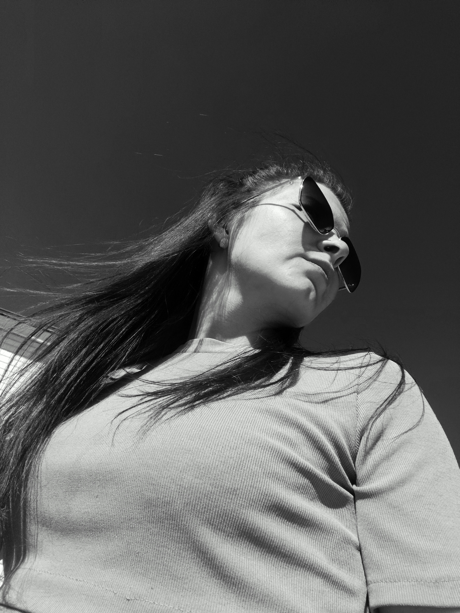 A low-angle, monochrome shot shows a person with long hair and sunglasses looking slightly downward. The subject is wearing a ribbed top. The background is a dark, plain sky. No specific location in Фоки, Russia, is discernible from the image.