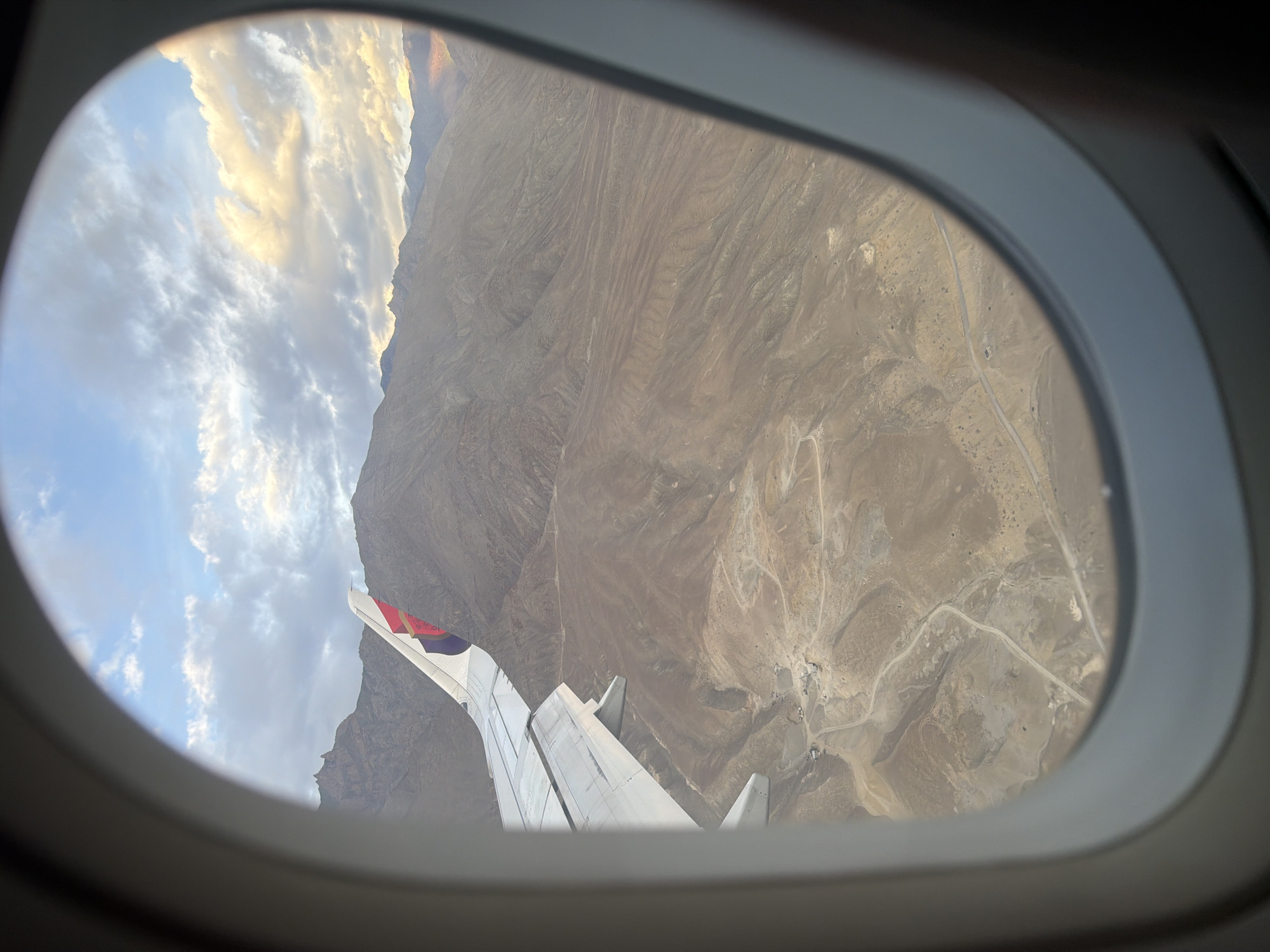 An aerial view from an airplane window shows a vast, arid mountain landscape. The foreground features the wing of the aircraft, partially obscuring the view. The terrain below is rugged and dry, with winding roads visible in the valley floor. There are some small clusters of buildings or structures scattered in the landscape, hinting at human settlement. The mountains in the distance are stark and rocky, with soft clouds and a warm glow in the sky, suggesting either sunrise or sunset. The scene appears to be over the region of Phey, India, characterized by its mountainous and arid geography.