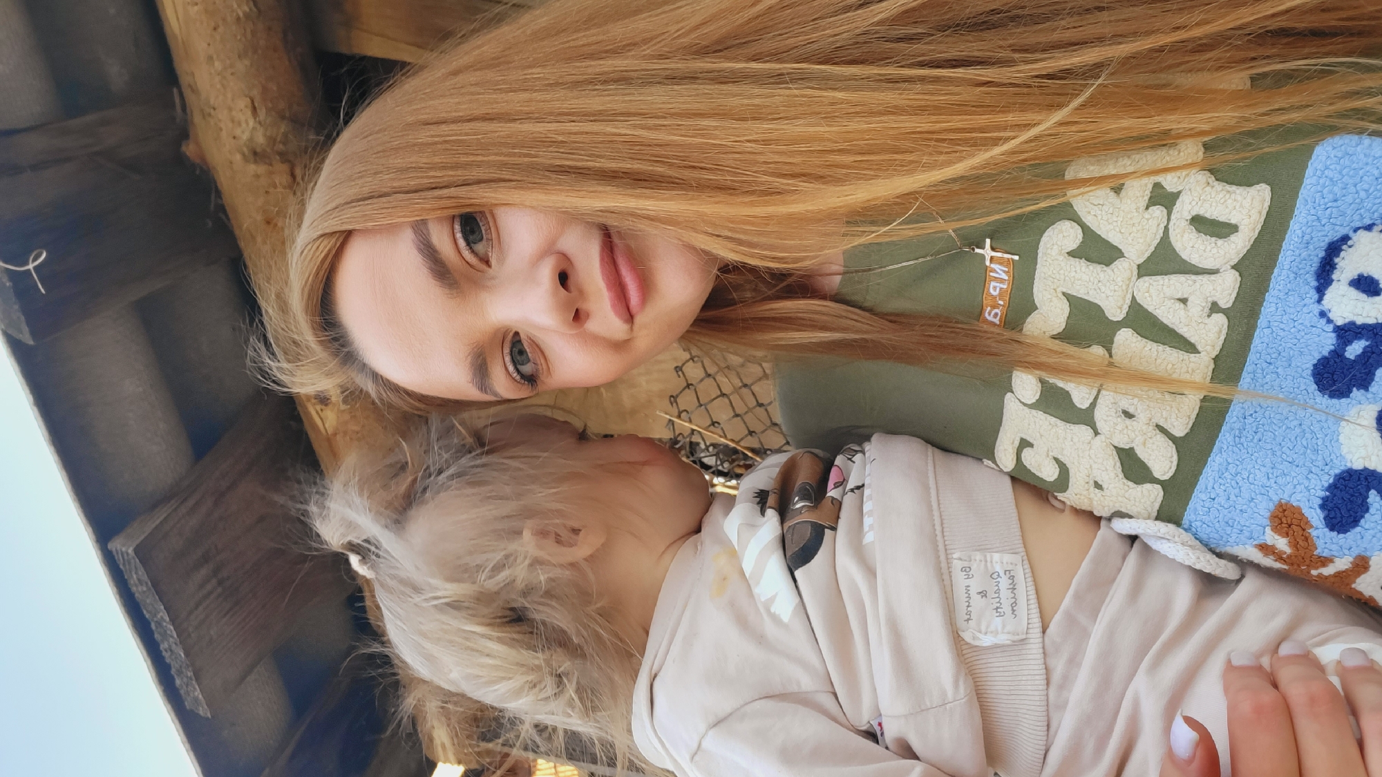 A low-angle, close-up shot shows a fair-skinned woman with long, straight, light-brown hair holding a fair-skinned toddler. The woman looks at the camera with green eyes and light pink lips. She wears a light green shirt with a white, blue, and brown design. The toddler, with wispy blonde hair pulled back, rests against the woman. The toddler wears a light-colored jacket and pants. A metal mesh fence sits behind the woman. Above them is a rough wooden structure. The location is Зипуново, Russia.