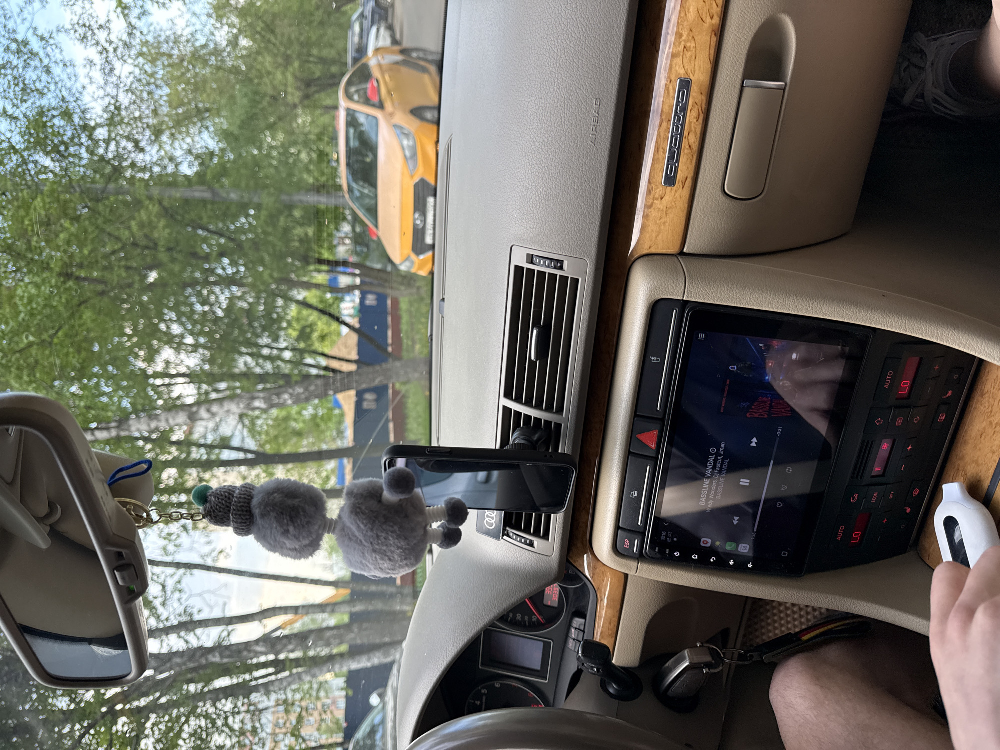 The image shows the interior of an Audi vehicle, likely an A4 B7 based on the dashboard design, with a view through the windshield. Outside, trees and buildings are visible, with a bright yellow car parked across the street. An accessory hangs from the rearview mirror. A smartphone is mounted on the dashboard in front of an air vent, the screen is black. The infotainment system displays music, and an electronic device is in the driver's hand to the right. Based on the location and cars, it is potentially Moscow, Russia.
