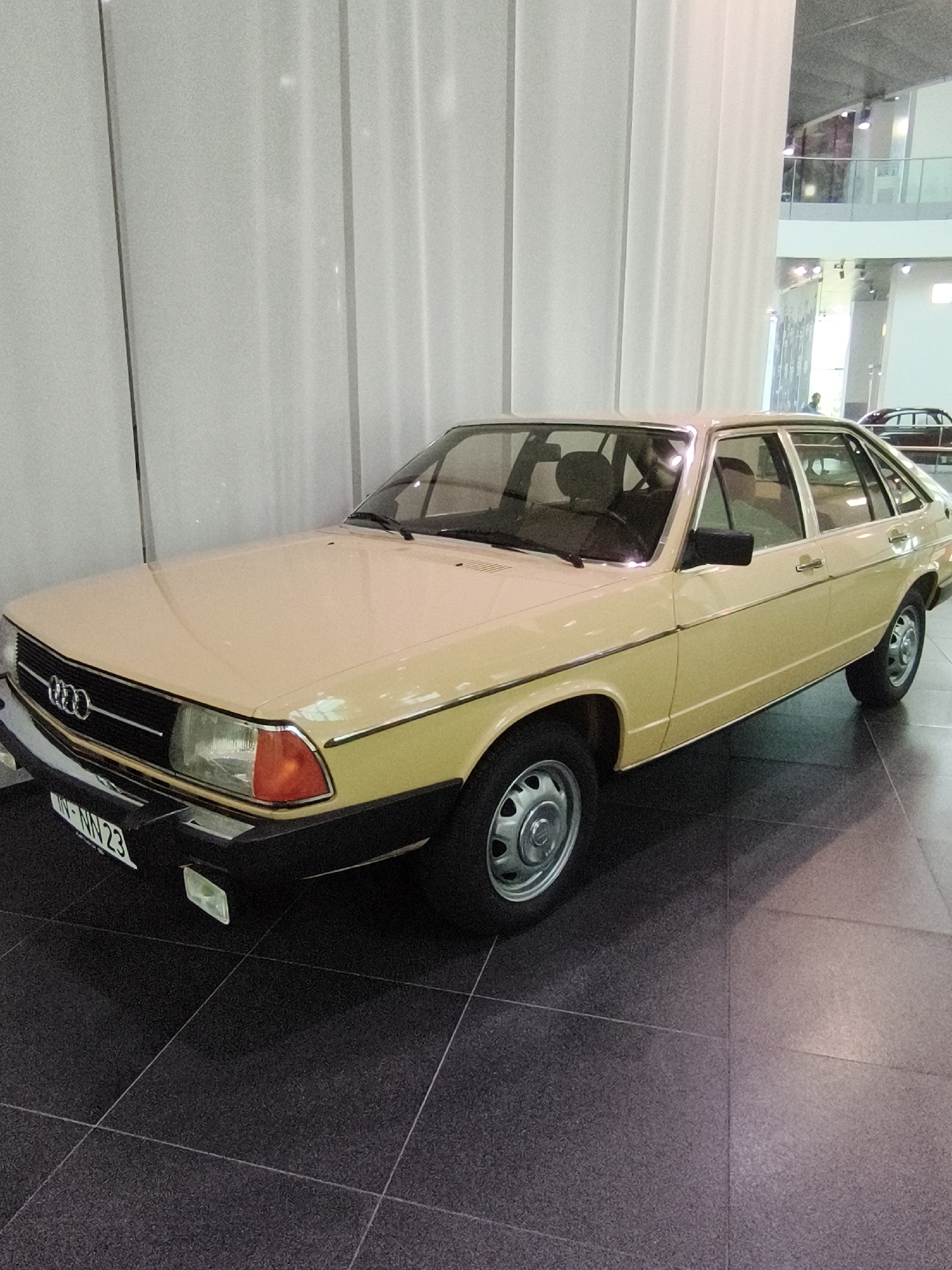 A light yellow Audi 100 (C2) is parked on a dark tiled floor. The car is a five-door hatchback, facing the viewer's left. The car's headlights are on. The license plate reads "IN-NN 23". The Audi rings emblem is visible on the grille. The car is in a large, well-lit indoor space with large vertical white fabric panels on the left side of the image. In the background, the upper levels of a modern building are visible, with glass railings and people. This scene appears to be inside the Audi Forum Ingolstadt museum in Ingolstadt, Germany, which houses a collection of Audi vehicles and historical exhibits.