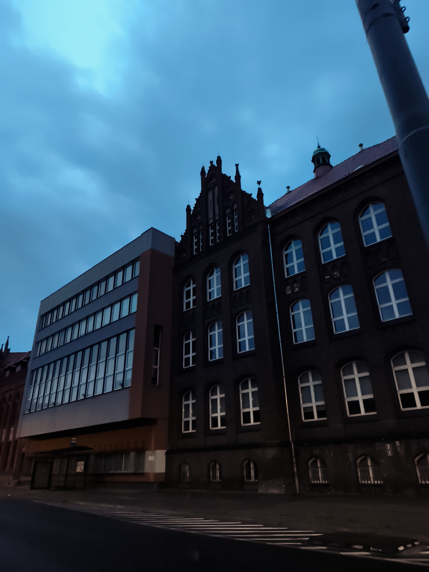 The image captures the exterior of a building complex in the city of Gdańsk, Poland, during twilight hours. The architecture suggests a combination of modern and older structures. On the left, a more contemporary building with a light-colored facade features multiple rows of rectangular windows, reflecting the dusky sky. Adjacent to it, a darker, older building showcases arched windows with white panes, characteristic of historical European architecture. The sky above is a deep, cloudy blue, indicating low light conditions. A street with crosswalk markings is visible in the foreground, with a portion of a dark lamppost or utility pole extending into the frame from the right edge. The scene appears to be devoid of people.