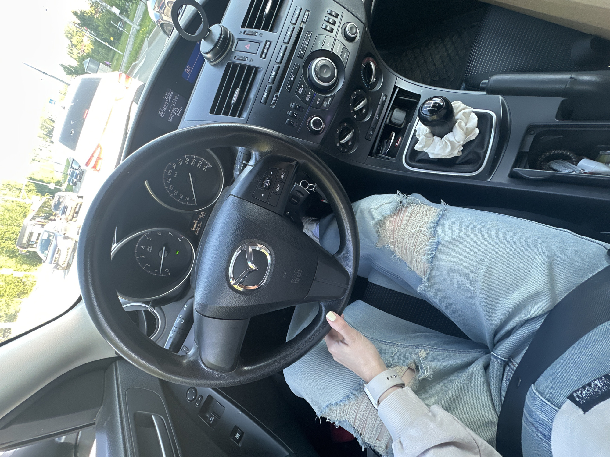 The image shows the interior of a Mazda car, viewed from the perspective of the driver. The driver's hands are on the steering wheel and shifter, and they are wearing ripped jeans and a light-colored sweater. The car is in motion, as indicated by the blurred background of trees and other vehicles. It is daytime, with bright sunlight. The location is the city of Perm, Russia. The dashboard displays a digital clock, temperature, and fuel efficiency. The gear shift has a scrunchie placed over it.