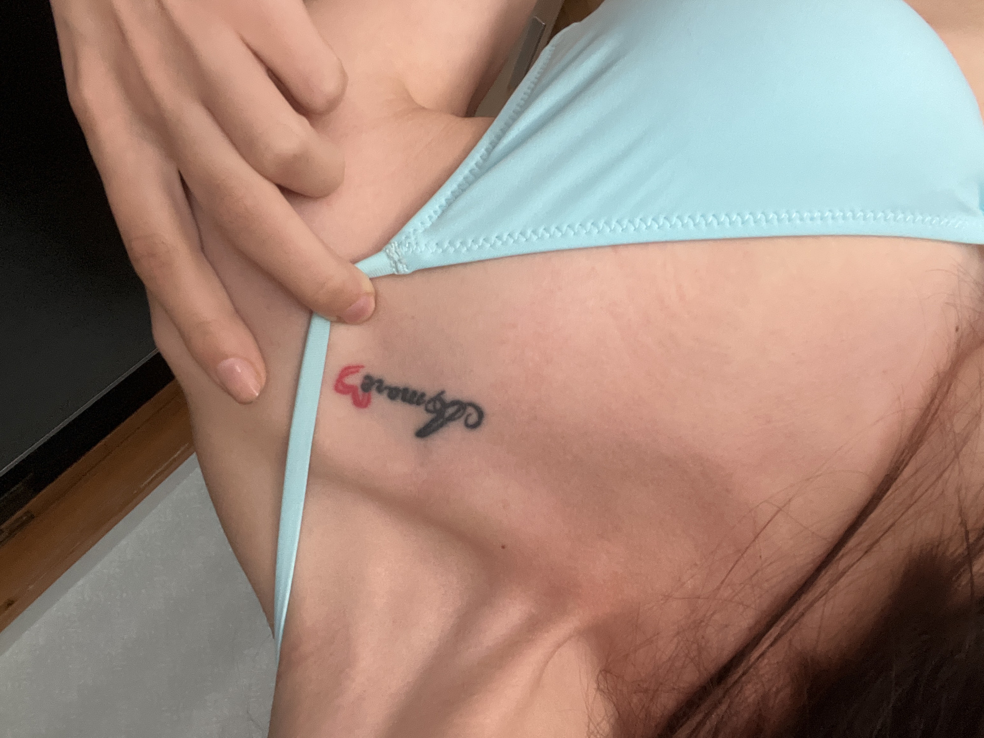 This is a close-up, vertical, eye-level shot of a person's collarbone and upper chest area, wearing a light blue bikini top. The skin tone is fair. There is a small, black tattoo on the left collarbone that appears to spell "Amore" with a red heart integrated into the design, specifically above the "o" and extending slightly to the right. A person's hand, with fair skin and light-colored fingernails, is visible at the top of the frame, with fingers gently resting on the shoulder and bikini strap. The background is blurred, with a pale wall on the left and a dark, wooden surface on the top right, suggesting an indoor setting. There is no information in the image to indicate the location is in Kyiv, Ukraine.