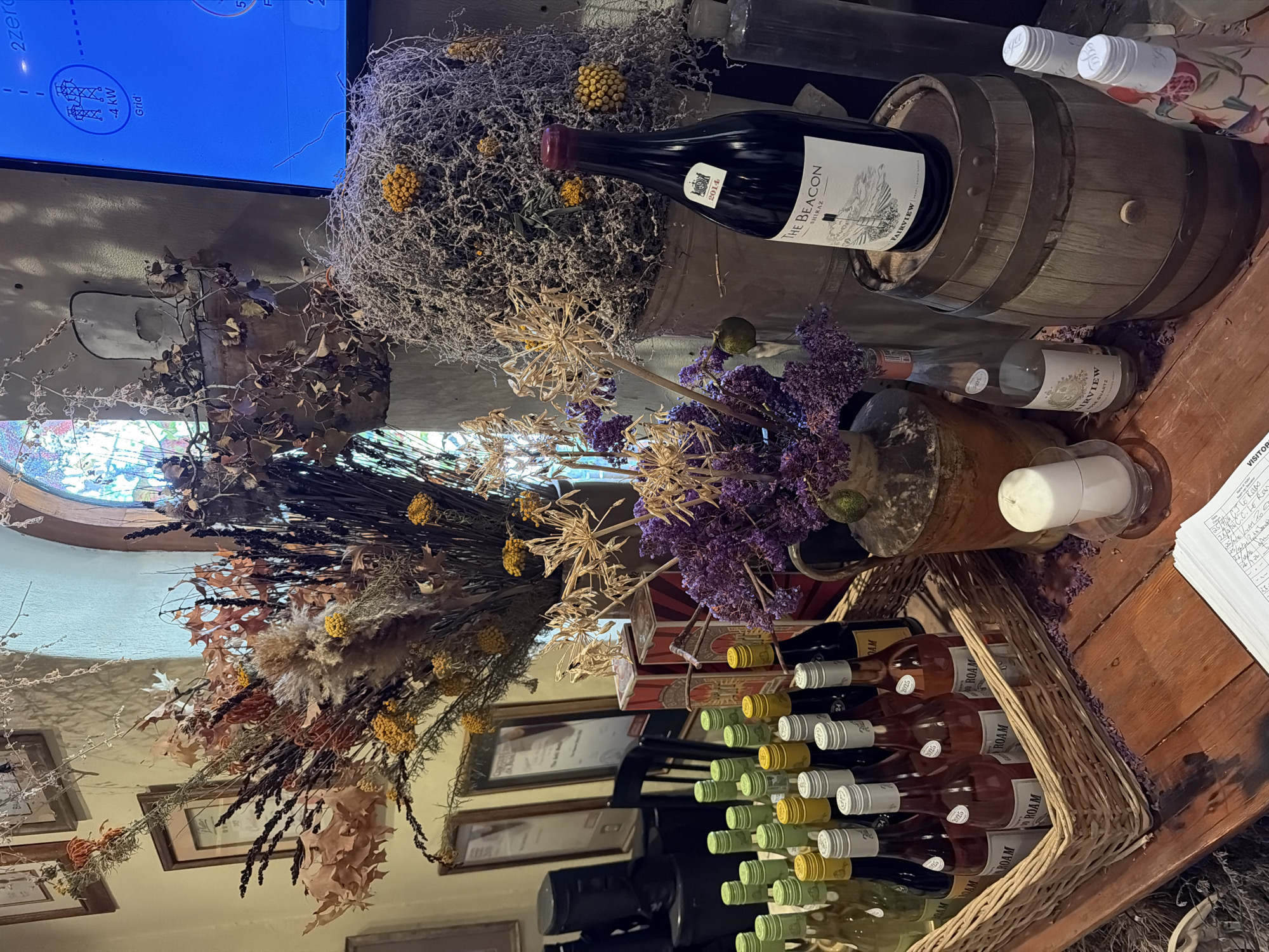 This image depicts a rustic and richly decorated interior scene, likely a wine tasting room, cellar, or shop, given the context of Drakenstein Nu, South Africa. The space is adorned with abundant dried foliage, including various grasses, leaves, and clusters of small yellow and purple flowers, creating a warm, autumnal aesthetic.
The central subjects are numerous wine bottles and wooden wine barrels. A prominent dark red wine bottle, labeled "THE BEACON SHIRAZ FAIRVIEW 2014," rests on its side atop a large wooden barrel. Below, a wicker basket is filled with multiple bottles, primarily labeled "ROAM," with light-colored labels and white caps, several also displaying "2025 AM." Another bottle, "FAIRVIEW QUARTZ," is also visible. The "Fairview" brand suggests a direct connection to the Fairview Wine and Cheese estate in the region.
In the background, a blue digital screen displays technical data, including "-4 kW Grid" and "2zero," juxtaposing modern technology with the rustic decor. The screen also shows text that appears to be "JAW GRIL." A rounded window with patterned or stained glass adds architectural detail. Other notable objects include framed pictures on the walls, a white candle in a holder, and a clipboard with a "VISITOR" sign and handwritten notes, further indicating a public or guest-oriented space. No people are visible, allowing the focus to remain on the curated display of wine and decorative elements.