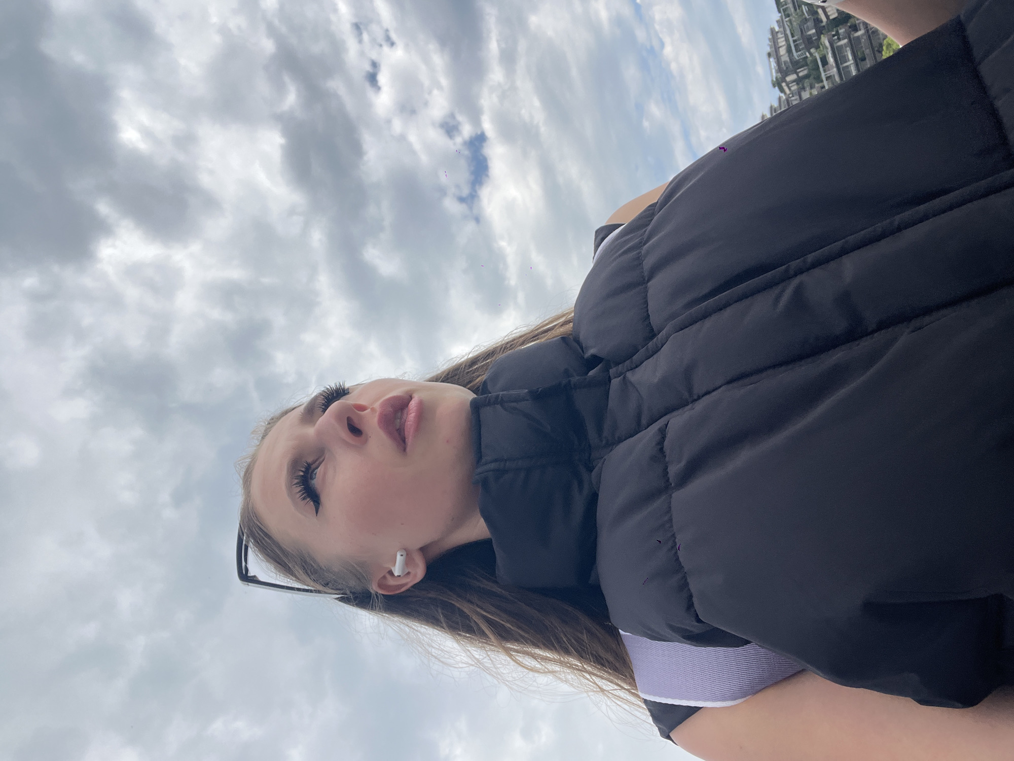 A young woman is captured in an outdoor setting in Budva, Montenegro, looking upwards at a cloudy sky. The image is taken from a low angle, emphasizing the sky and her upward gaze. She has long, light brown hair, which falls over her shoulder, and prominent dark eyelashes. Her lips are slightly parted, revealing a light pink hue. She wears a white wireless earbud in her right ear and black sunglasses are pushed up onto her head, resting in her hair. She is dressed in a black sleeveless puffer vest, with a white and light purple striped top visible underneath on her right side. The sky is largely overcast with shades of grey clouds, though hints of blue are visible in some areas. In the upper right corner of the image, behind her, a cluster of modern-looking buildings with green foliage is visible, suggesting an urban or resort environment. The overall impression is a candid, relaxed moment on a cloudy day. There is no visible text in the image.