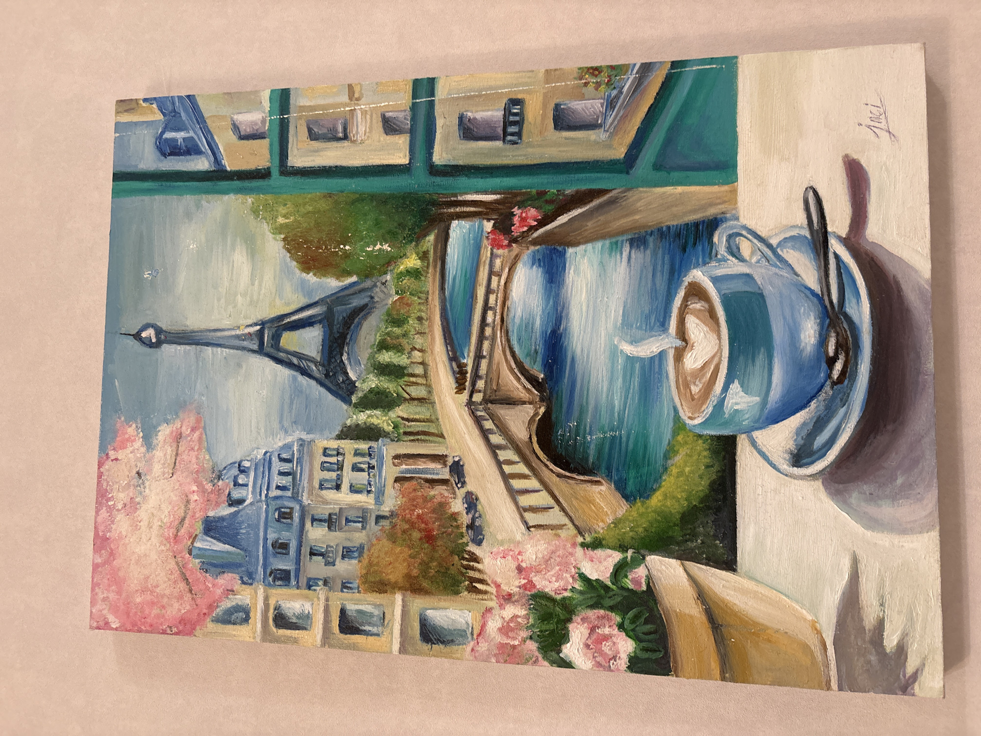 This is an oil painting depicting a scene in Paris, France. In the foreground, a cup of coffee with a heart-shaped latte art sits on a saucer next to a spoon. To the left, a potted plant overflows with pink flowers. The background features a picturesque view of the Eiffel Tower, a river with a bridge, and classic Parisian buildings, some with mansard roofs and balconies. Trees with fall foliage add color to the scene, and cherry blossoms are visible on the left. The painting has a textured appearance, characteristic of oil paints. The artist's signature, "Joci," is visible in the bottom right corner. This scene is not located in Baku, Azerbaijan, but rather in Paris, France.