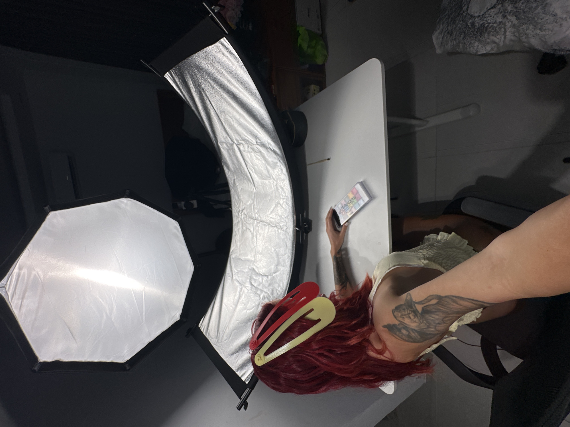 The media file shows a person with vibrant red hair, adorned with large red and yellow hair clips. They are wearing a light-colored top with ruffles around the neckline. The person has tattoos visible on their arm and neck. They are holding a small object, possibly a phone or a card, in their right hand. The scene appears to be a home studio setup, with professional lighting equipment visible. A large, octagonal softbox with a diffused light source is positioned to the left of the frame. Another lighting modifier, a silver reflective strip box, curves around the person from the top left. The background is dimly lit, with some shelves and objects visible in the distance, suggesting an indoor setting. The overall lighting suggests an artificial light setup, likely for photography or videography. The image is oriented vertically, with the person's head towards the bottom of the frame and their legs towards the top. There is no visible information about the time of day, weather, or specific emotions conveyed by the subject. There is some text on the object being held by the person, but it is not legible. The context provided indicates the location is North Kuta, Indonesia.