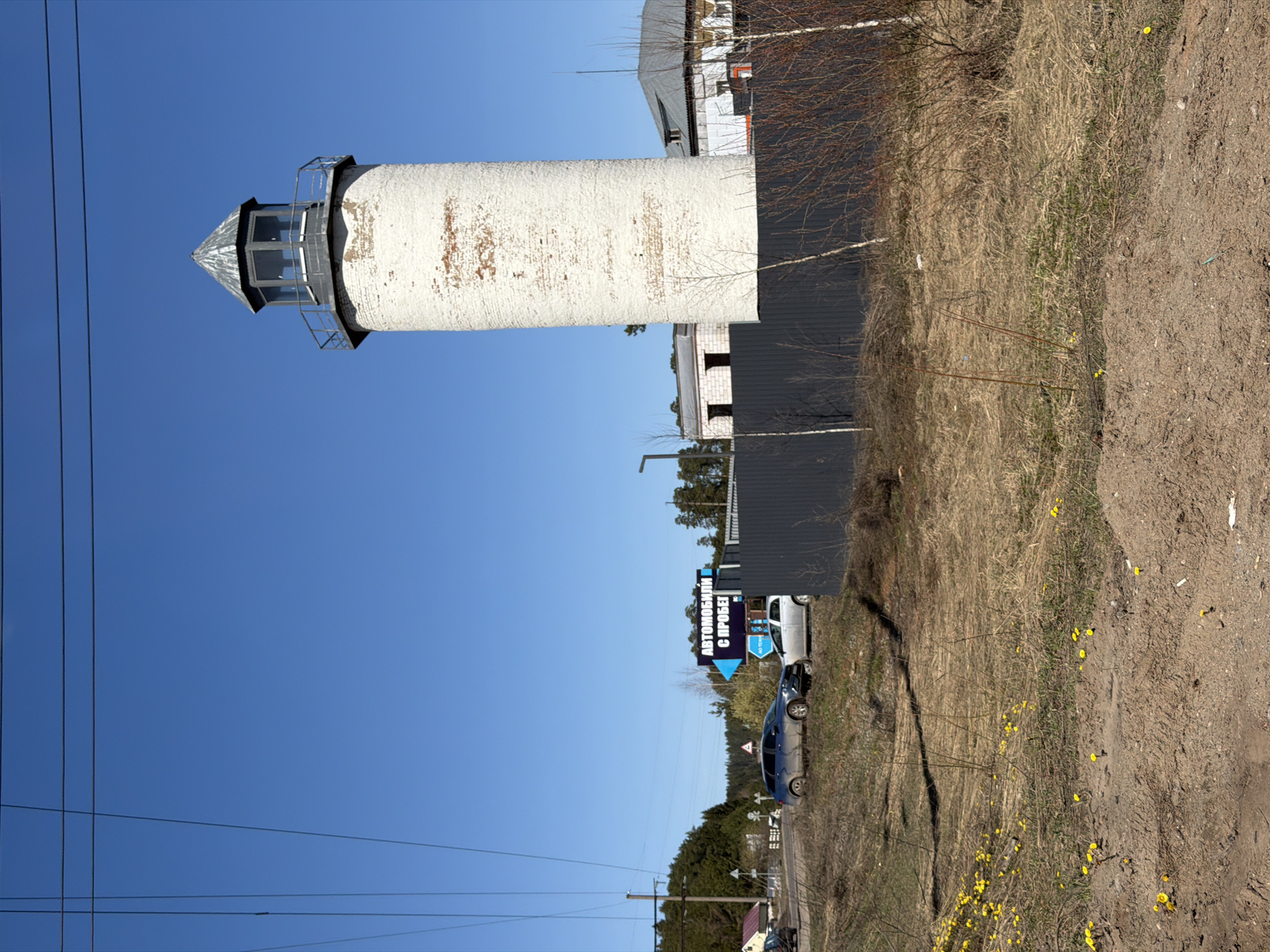 The image shows a white cylindrical structure with a dome-shaped top, possibly a lighthouse, in Votkinsk, Russia. The structure is adjacent to a building with a dark grey facade, visible behind some sparse trees. Next to the building, there's a road with parked cars and a sign in Russian that reads "АВТОМОБИЛИ С ПРОБЕГОМ," which translates to "USED CARS." Power lines traverse the blue sky in the upper-left quadrant of the image. The ground is covered with dried grass and patches of bare soil with some yellow flowers.