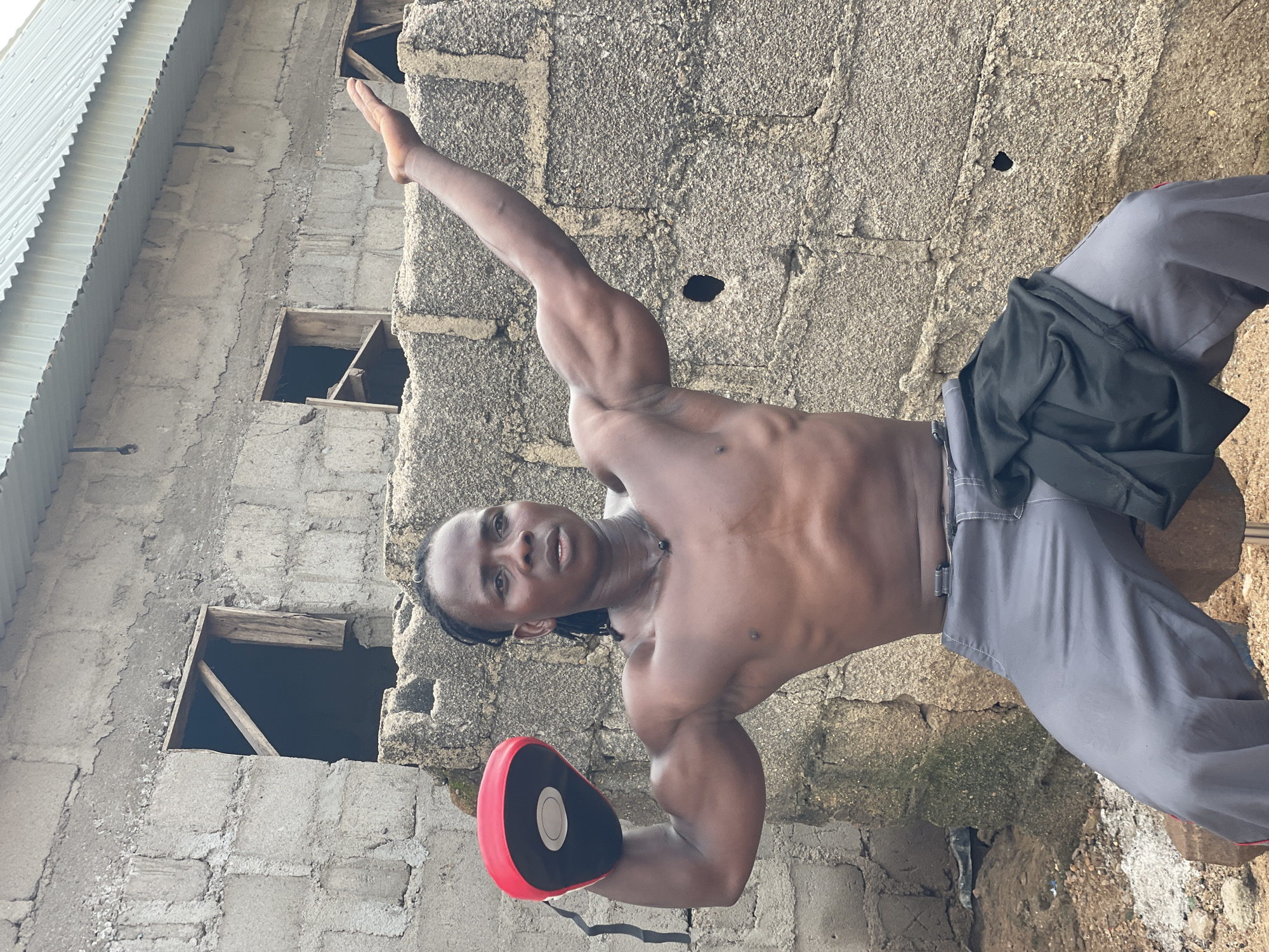 A shirtless young man with braided hair and a necklace is posing outdoors in front of a rough, gray cinder block building, possibly in Akwanga, Nigeria. He is wearing gray pants and a black garment is draped over his left thigh. He holds a red and black boxing pad in his left hand, and his right arm is raised, palm facing forward. The building has several openings, some with wooden frames. The ground is dirt and scattered with debris.