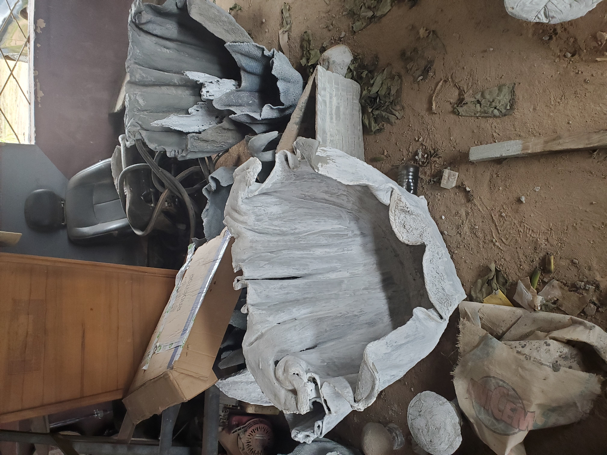 The image depicts an indoor storage area, possibly a workshop or garage, in Jos, Nigeria. The scene is cluttered with various objects, including large, irregularly shaped concrete planters, a discarded car seat with a steering wheel, a wooden cabinet or desk, and a cardboard box with visible text indicating "RADIATOR". The floor is covered in dirt and debris, with scattered dried leaves. In the foreground, a burlap sack with visible red lettering is partially visible, along with other miscellaneous items. The overall impression is one of an unorganized and possibly abandoned or under-construction space.
