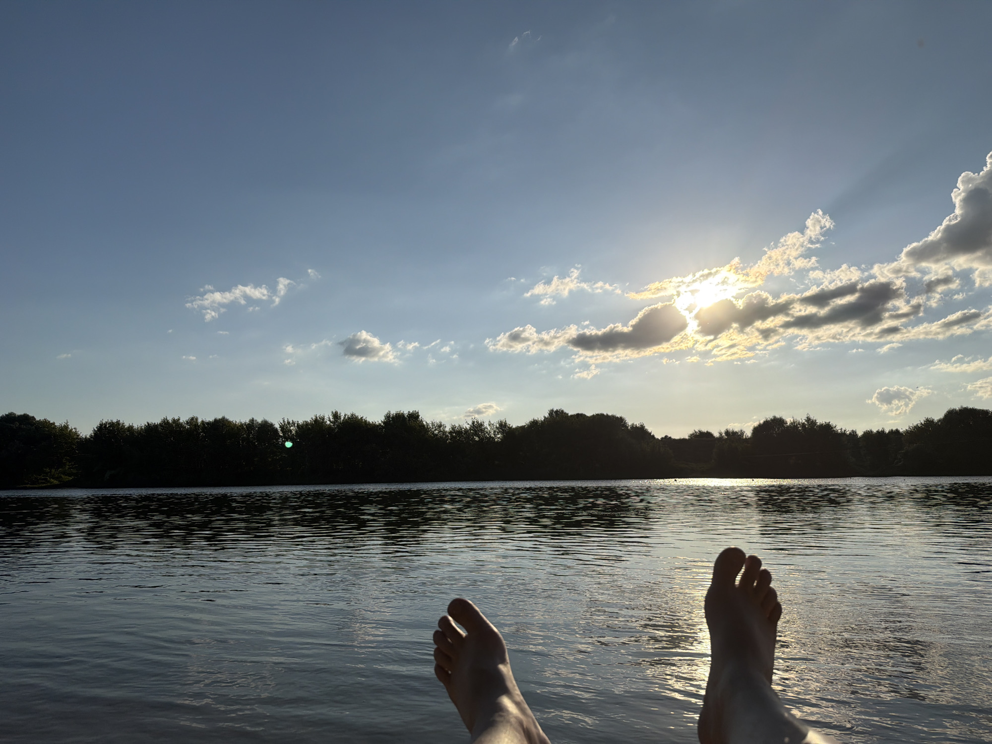 This image depicts a serene natural scene, likely at dusk, overlooking a body of water. The foreground features a pair of bare feet resting, suggesting a relaxed posture. The middle ground is dominated by the calm surface of a lake or river, reflecting the sky and surrounding trees. In the background, a dense line of dark green trees forms a silhouette against the horizon. The sky is a gradient of blue, with scattered white and gray clouds. The sun is setting, creating a bright, hazy glow behind a cluster of clouds, casting rays of light across the sky and water. A small, bright green circular light is visible near the center of the treeline on the horizon. The overall impression is one of tranquility and natural beauty. The location is identified as Скорняково, Russia.