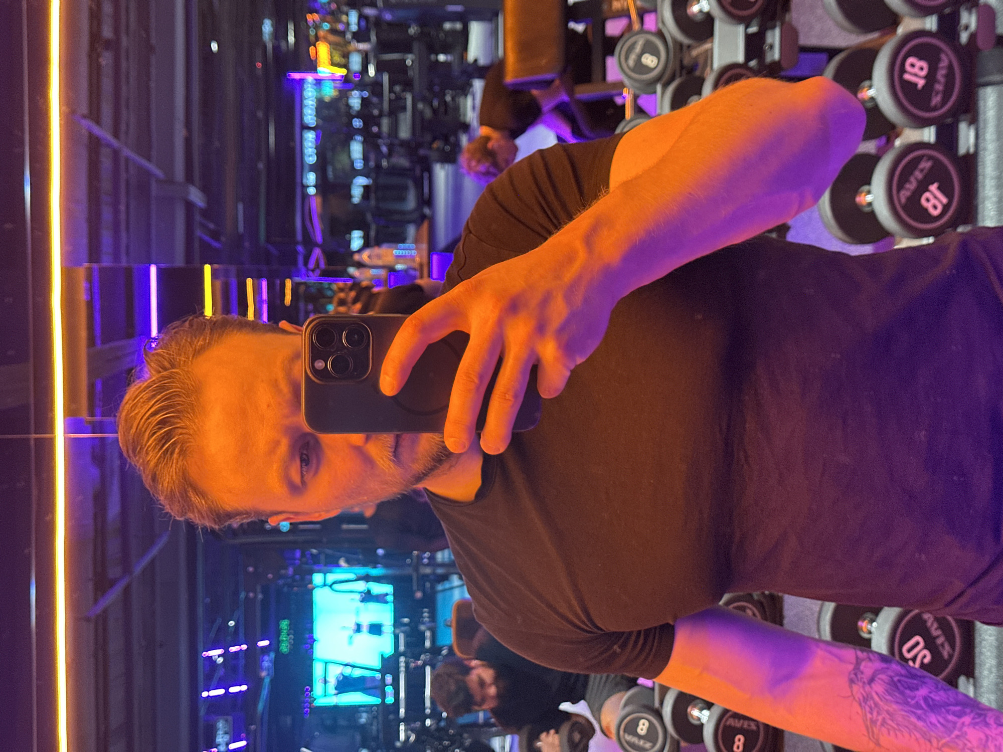 The image shows a man taking a selfie in a gym. The man has blonde hair and is wearing a black t-shirt. He is holding a black phone in his right hand, partially obscuring his face. His left arm is visible, with a tattoo on the forearm. In the background, there are rows of dumbbells and weight machines. Other people are present in the gym, seemingly working out. There are bright neon lights in the gym, creating a colorful atmosphere. The location cannot be determined to be Sochi, Russia from the given image.