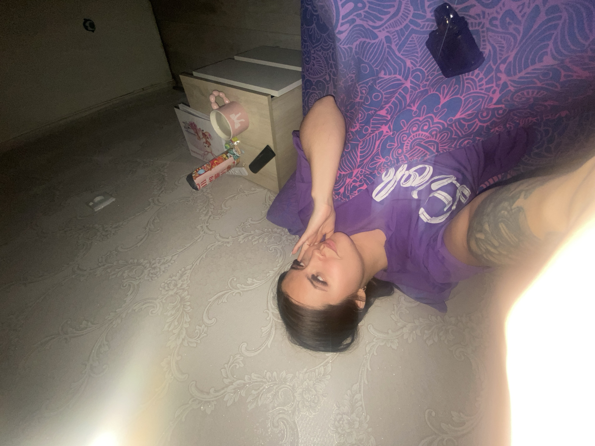 A woman with dark hair is lying on a patterned, possibly wallpapered, floor in a dimly lit room. She is wearing a purple t-shirt with white writing. Part of her arm, which has a tattoo, is visible. Next to her is a nightstand with a pink mug, a book, and a small black object. A patterned purple blanket or sheet is draped behind her. The location appears to be indoors, potentially in a private residence. The image was taken from an overhead perspective. The location in the city of Chaikovsky, Russia cannot be determined from the image.