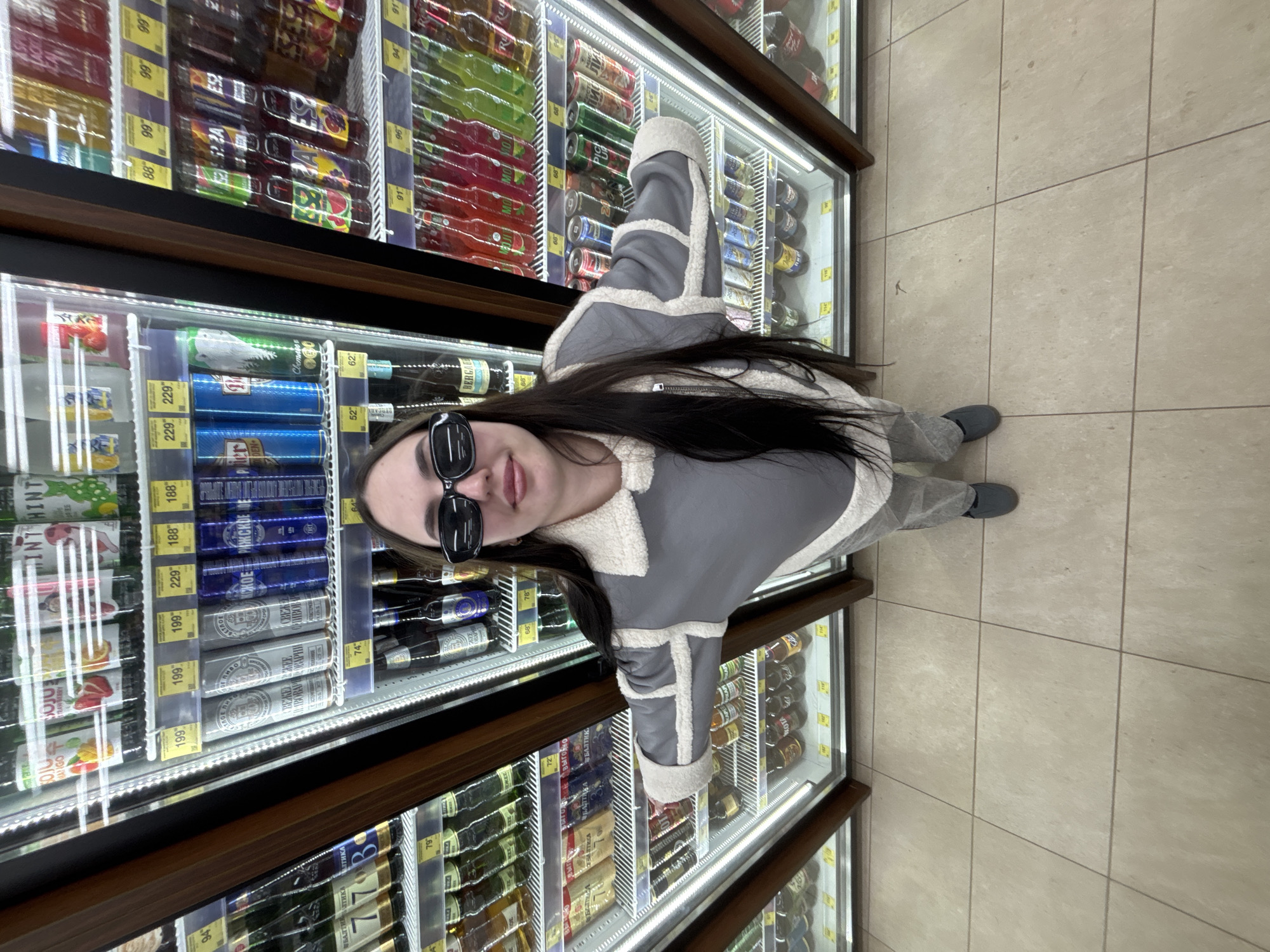 An overhead, slightly angled shot captures a young woman standing in a supermarket aisle in what appears to be Фоки, Russia. She has long, dark hair and is wearing sunglasses, a grey and cream faux-fur lined jacket or coat, grey trousers, and dark slip-on shoes. She is looking upwards towards the camera with a subtle smile.
The woman is positioned between two long, brightly lit refrigerated display cases, which are filled with an assortment of canned and bottled beverages. The products include various soft drinks, juices, and alcoholic beverages such as beer. Many items have visible price tags and labels featuring Cyrillic text.
Notable text includes brand names like "OJU MANGO," "OJUS STRAWBERRY," "HINT HINT," "БАЛТИКА" (Baltika), "СВЕТЛОВ," "РИЖСКОЕ," and "BERGAB." Several items are clearly labeled as "ПИВО" (beer) or "СВЕЖЕЕ ПИВО" (fresh beer). Prices are prominently displayed on yellow and white tags, ranging from approximately 52 to 229, likely in rubles. The floor is covered with large, light-colored square tiles. The setting suggests an everyday shopping experience.