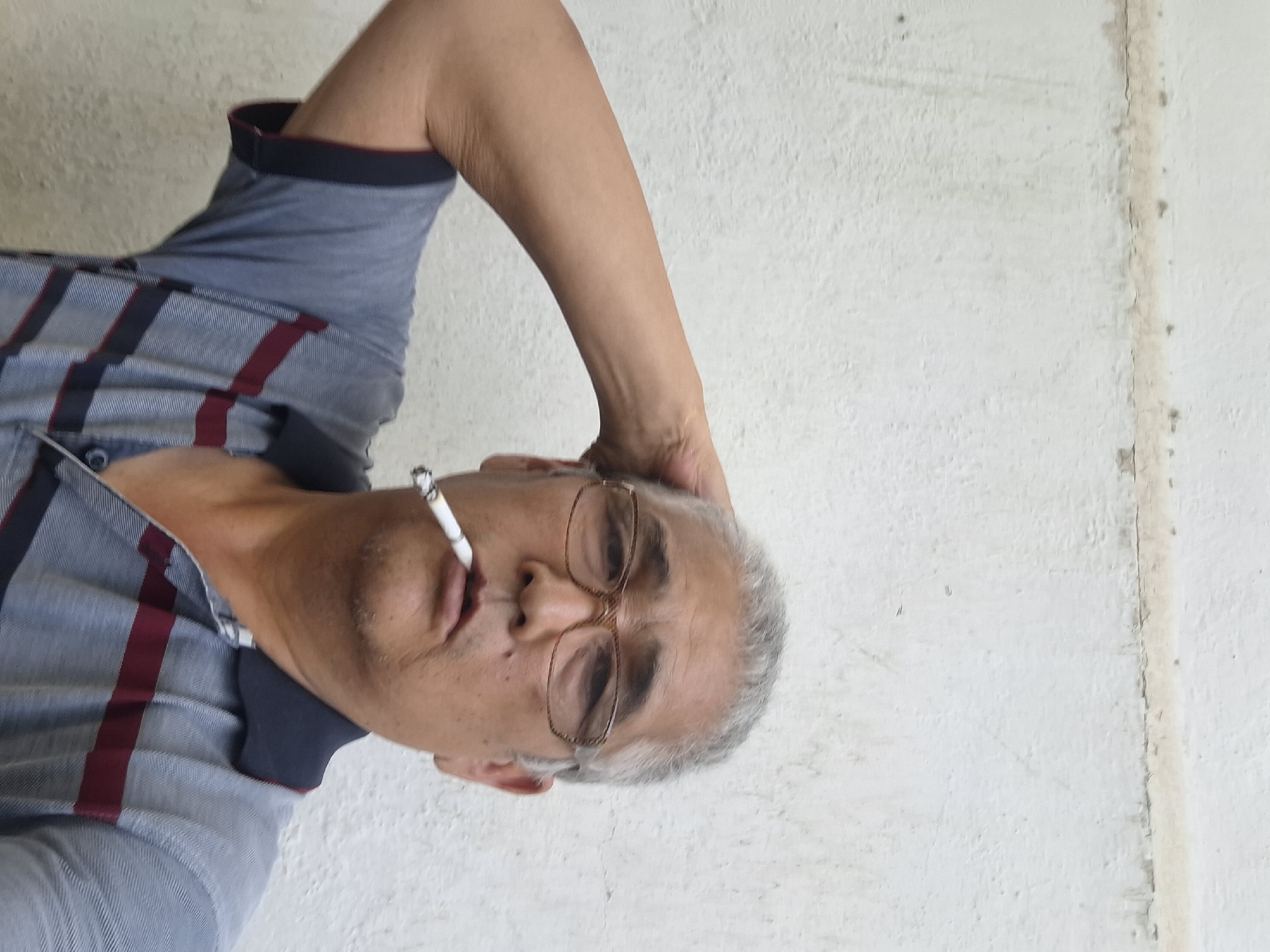 A middle-aged East Asian man stands with his right arm raised and hand on his head, looking directly at the camera. He is wearing a grey polo shirt with navy and maroon stripes. A cigarette is held in his mouth. He has short, grey hair and is wearing glasses. The background is a textured, off-white wall. The scene appears to be outdoors, with natural lighting. There are no specific landmarks visible to indicate a location within the city of Almaty, Kazakhstan.