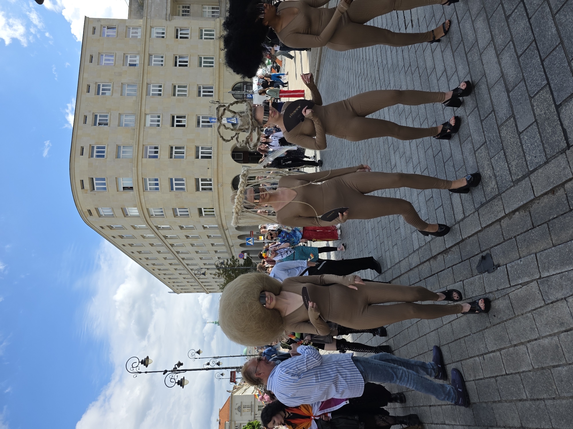 The image depicts a scene in Warsaw, Poland, likely during a parade or festival. In the foreground, four individuals are walking on a cobblestone street. The central figure is a woman with an exceptionally large, spherical, light brown wig and oversized, reflective sunglasses. She is wearing a form-fitting, tan-colored jumpsuit and holding a dark fan. To her left and right are two other women in similar tan jumpsuits and distinctive, elaborate headwear. The woman to the left has a woven, cage-like headdress, and the woman to her right has a braided, sculptural headdress. The fourth individual, on the far right, is also wearing a tan jumpsuit and has a large, dark, afro-style wig. Behind these individuals, other people are visible, some holding rainbow flags, suggesting a Pride event. In the background, a large, pale, multi-story building with numerous windows curves around a corner. Streetlights and other architectural elements are also present. The sky is blue with scattered white clouds.