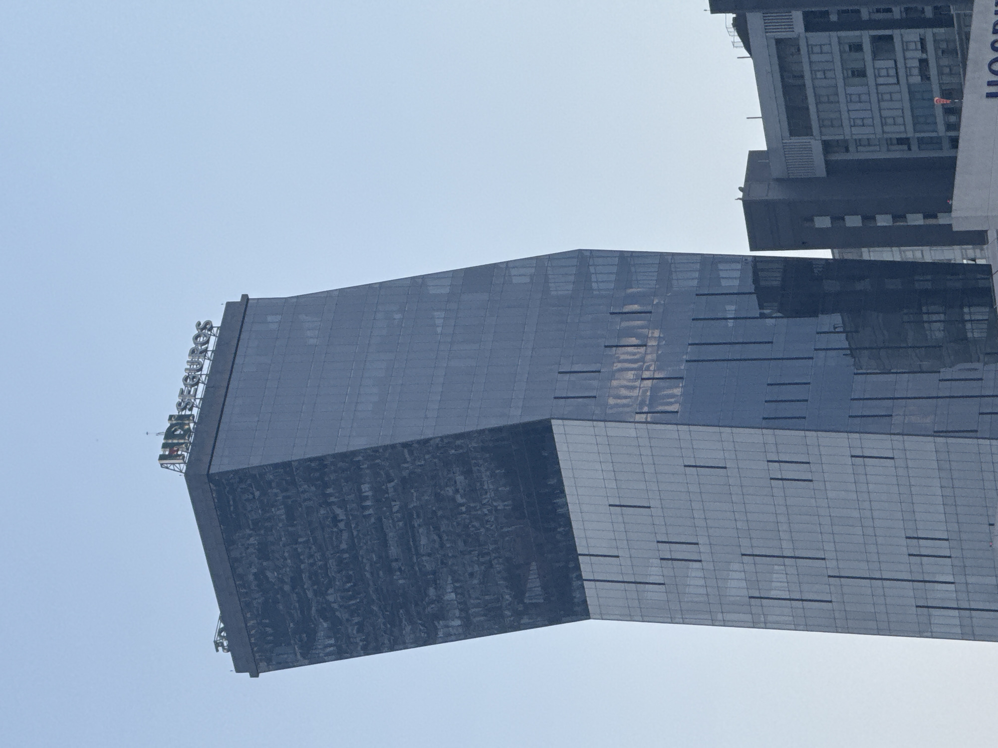 A low-angle shot shows a tall, modern glass building that occupies most of the frame, with its upper section slightly angled to the left. The top of the building features the word "HERSH" in large letters. The building's facade is largely reflective, with a pattern of horizontal lines. Adjacent to the main structure is another, taller building with a visible grid pattern of windows and what appears to be a partial view of the word "HOSPIT". The sky is a pale blue, providing a neutral background.