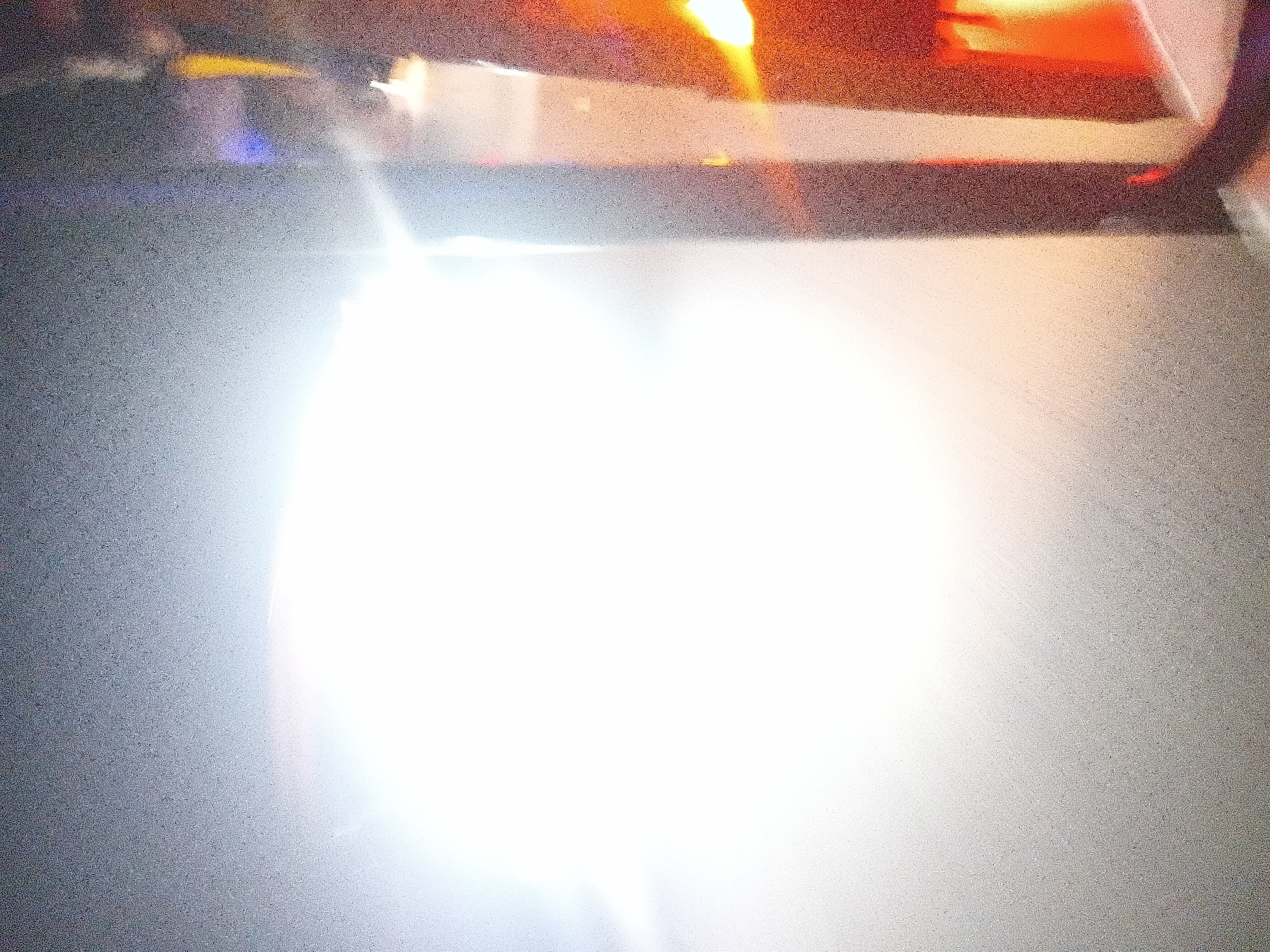 The image depicts a dimly lit interior space with a bright, diffused light source dominating the left side of the frame, creating a lens flare effect. The right side of the image shows vertical structures with varying colors, including dark brown, orange, and possibly some blue and yellow hues, suggesting architectural elements or furniture. The texture of the image is grainy, indicating low light conditions or a specific photographic technique. Without further context or visual cues, it is impossible to definitively identify the location as Jalingo, Nigeria, or to provide specific details about people, objects, or actions. The scene appears to be indoors, possibly a performance venue, exhibition space, or a room with ambient lighting.
