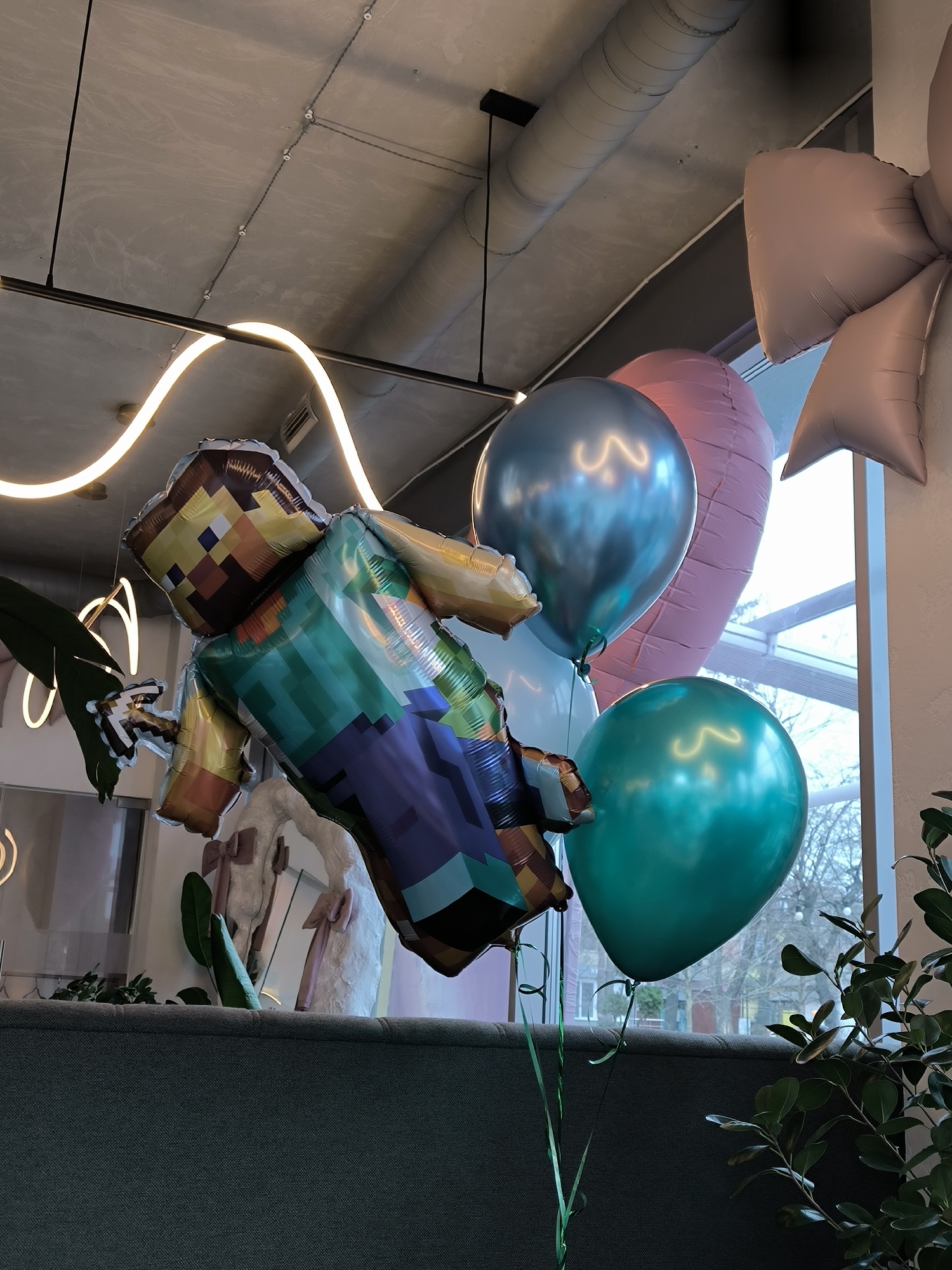 The image shows a cluster of celebratory balloons, including a large Minecraft character balloon, in an indoor setting. Other balloons include a silver, a pink oblong, and a teal-colored round balloon. The balloons are tethered with green ribbons.
The location is identifiable as Vinnytsia, Ukraine, based on the view through the window in the background. The exterior scene shows bare trees and buildings typical of a city environment, consistent with the urban landscape of Vinnytsia.
The indoor setting appears to be a restaurant or event space. A grey upholstered sofa is visible in the foreground. In the background, there is a window, and a decorative white swan figure. Additionally, there is a contemporary lighting fixture on the ceiling. The environment suggests a festive or celebratory occasion.