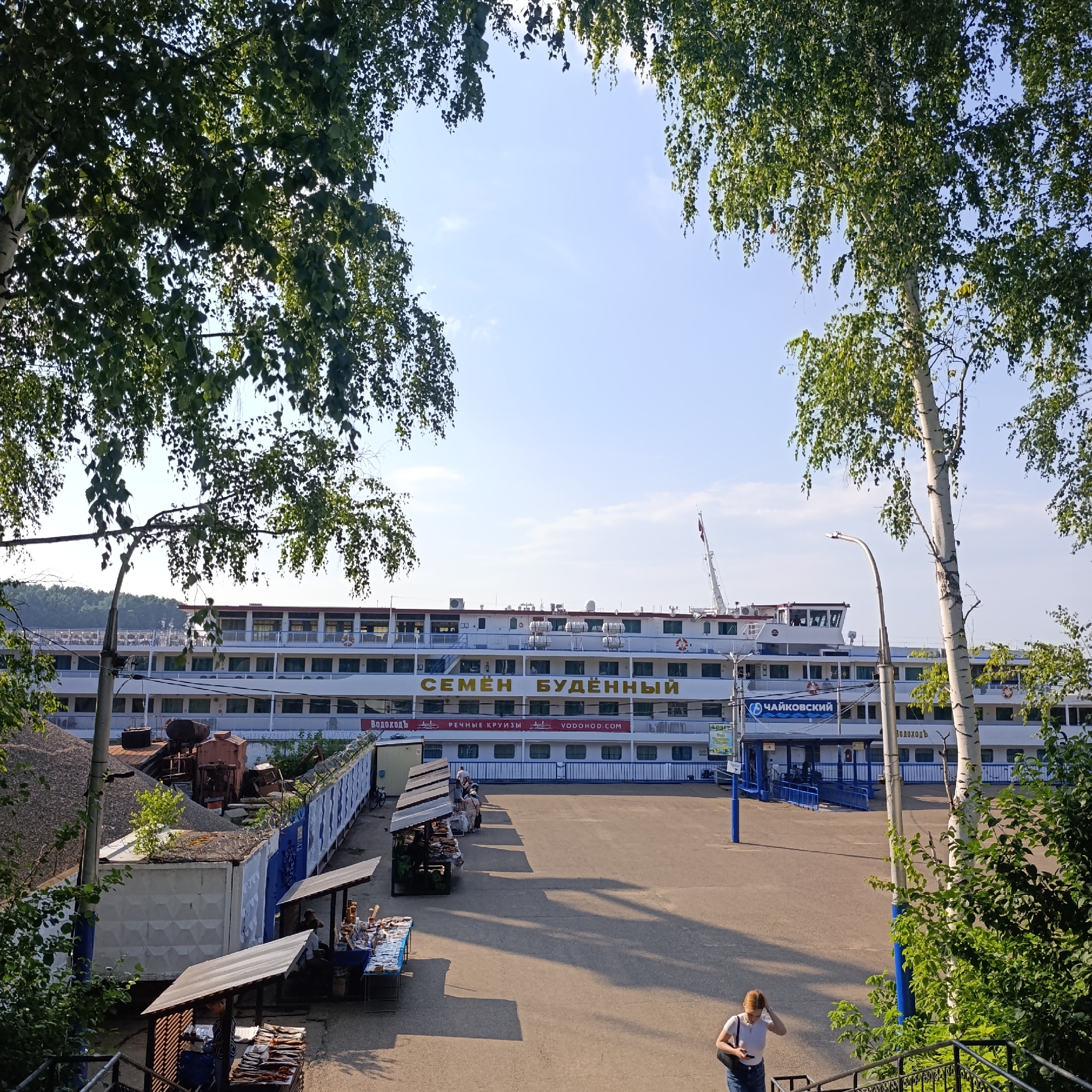 The image depicts a sunny daytime scene along the riverfront in Chaikovsky, Russia. Dominating the background is a large white river cruise ship named "СЕМЁН БУДЁННЫЙ" (Semyon Budyonny), with the name also visible in golden lettering. Another ship, "ЧАЙКОВСКИЙ" (Chaikovsky), is partially visible to the right. The ship is docked at a pier, with a blue ramp leading up to its entrance. Along the waterfront, there are market stalls with awnings, where people are seen browsing and selling goods. In the foreground, to the right, a woman in a white t-shirt and jeans is walking down a set of stairs, holding a dark bag and looking at her phone. Several birch trees with green foliage frame the top of the image, with sunlight filtering through their leaves. The ground in front of the ship appears to be a paved area, with long shadows cast by the market stalls. A barbed-wire fence runs along the edge of the pier area, separating it from a pile of gravel and some industrial equipment. The sky is clear blue with a few wispy clouds.