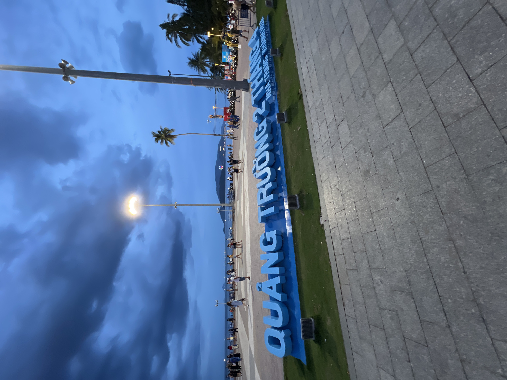 The image depicts a public square at dusk in Nha Trang, Vietnam, likely near the beach due to the presence of palm trees and a body of water in the background. Dominating the foreground are large, bright blue three-dimensional letters spelling out "QUẢNG TRƯỜNG 2 THÁNG 4 NHATRANG KHANH HOA". The letters are set on a green, grassy area bordered by a grey, tiled walkway.
In the midground, a paved promenade is visible, populated by numerous people strolling, standing, and gathering. They are casually dressed, suggesting a leisurely atmosphere. The background shows a body of water, possibly the ocean, with a distant landmass or island. The sky is a deep blue, characteristic of twilight, with dramatic, dark clouds overhead and illuminated by streetlights.
Several lampposts are strategically placed throughout the scene, some with multiple light fixtures, illuminating the area. One tall lamppost with speakers is prominent on the right side of the image. Other objects include what appear to be low-lying electrical boxes or lighting fixtures at the base of the large letters. The overall impression is a vibrant public space in the evening, likely a popular tourist destination or local gathering spot.
