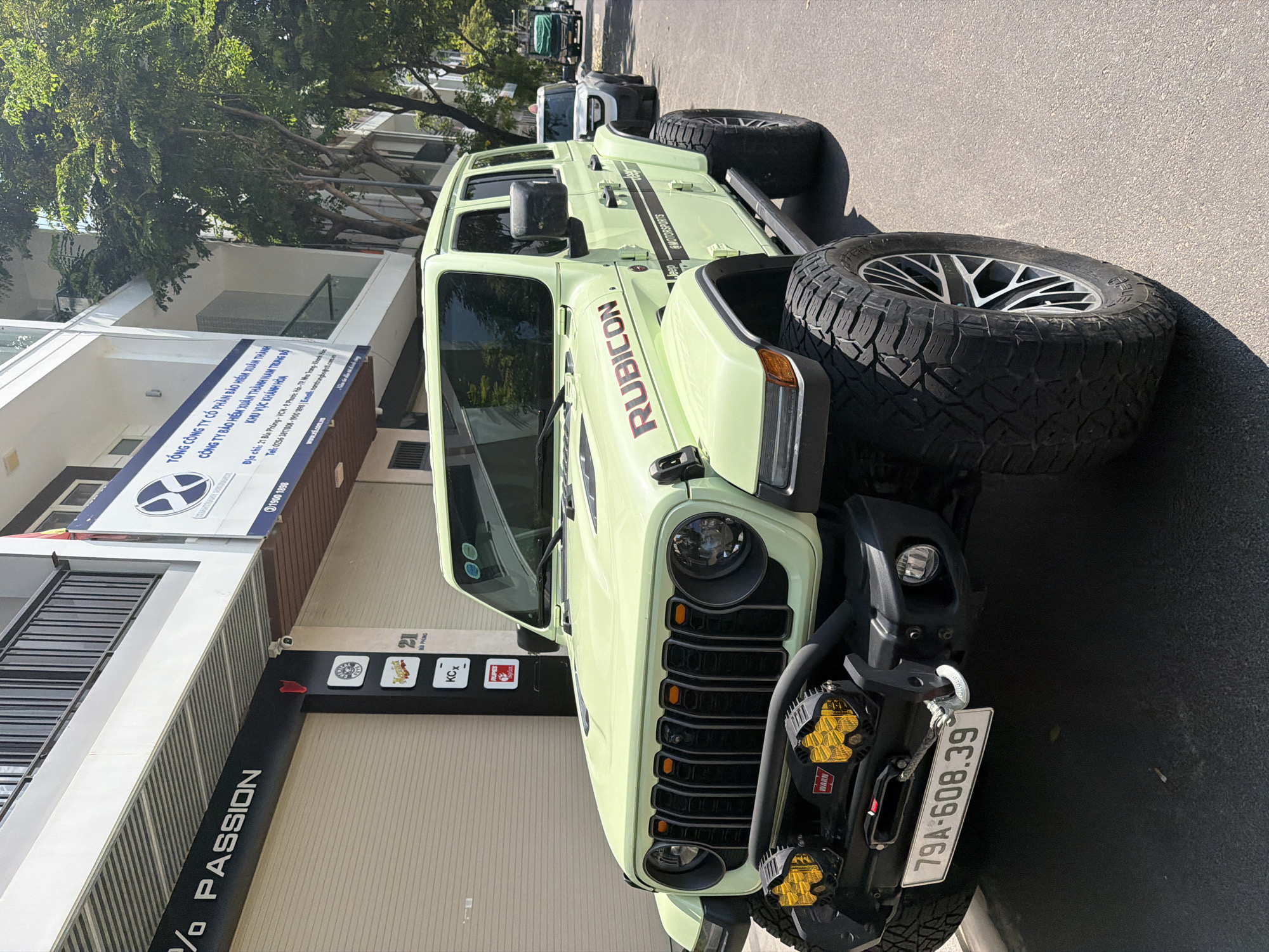 In Nha Trang, Vietnam, a light green Jeep Wrangler Rubicon is parked on the street in front of a building with a black awning that reads "% PASSION". The Jeep has large, aggressive off-road tires, a black grille with orange accents, and two yellow off-road lights mounted on the front bumper. A winch is visible behind the bumper. The license plate reads "79A-608.39". To the right of the Jeep, another white vehicle is parked, and further down the street, a green truck is visible. The building behind the Jeep has several signs, including one that says "TONG CONG TY CO PHAN BAO HIEM XUAN THANH TRONG BO". The environment is sunny with trees lining the street.