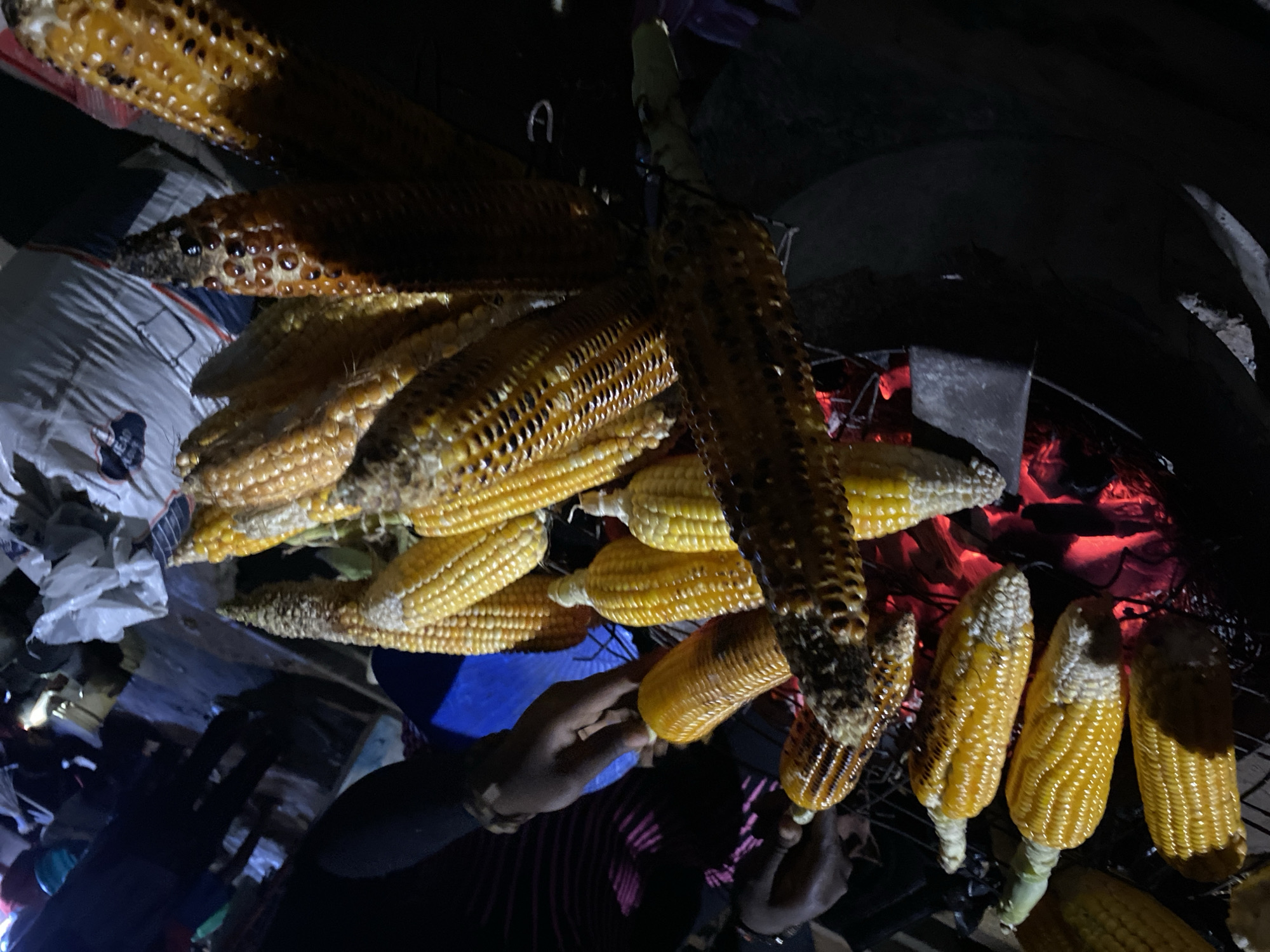 The image depicts a scene in Bukuru, Nigeria, featuring corn on the cob being roasted over a charcoal grill. Several cobs, some with visible charring, are arranged on a metal grill above glowing red embers. Hands are visible near the corn, possibly turning or tending to it. Background elements include bags of supplies in a crowded environment with dark shadows suggesting the scene is occurring at night.