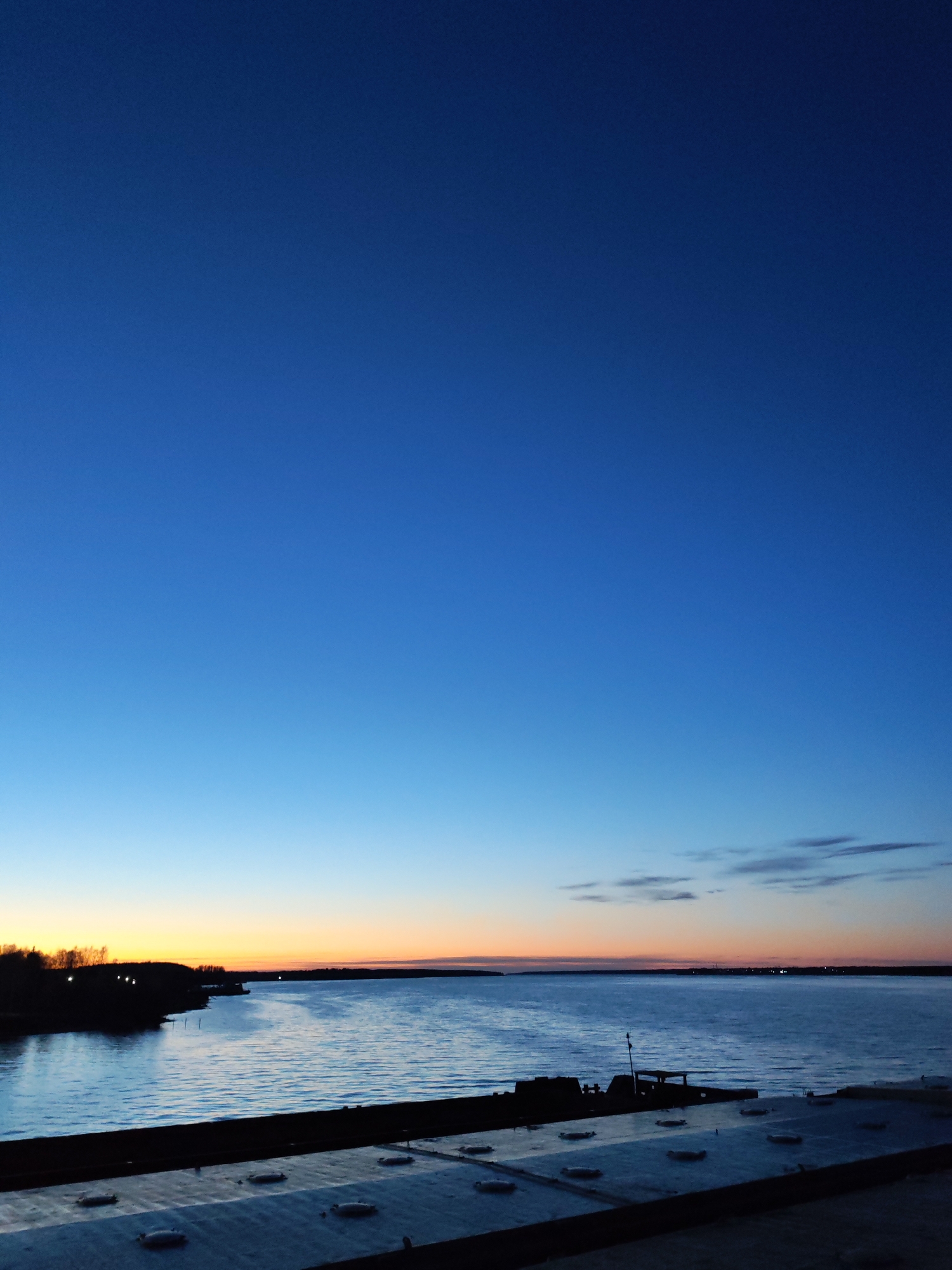 This vertical shot captures a tranquil twilight scene over a large body of water, likely a river or lake, during either dawn or dusk. The sky dominates the upper two-thirds of the frame, showcasing a dramatic gradient of blues. It transitions from a deep, dark royal blue at the very top, gradually lightening to a vibrant medium blue, and then to a pale sky blue closer to the horizon. Near the horizon line, a striking band of orange and yellow light indicates the setting or rising sun, casting a warm glow.
The water surface below reflects some of the sky's blue hues, appearing slightly rippled and calm. On the left side of the water, a dark, silhouetted landmass with trees is visible, dotted with a few faint, distant lights. Another similar dark shoreline stretches across the far horizon on the right. A few wispy, dark clouds are scattered in the lighter blue sky just above the distant horizon on the right.
In the immediate foreground, a dark, indistinct structure takes up the bottom portion of the image. It appears to be a flat, metallic or concrete surface, possibly part of a dock, pier, or bridge, featuring several circular, raised elements that resemble covers or vents. To the right, another dark, angular structure is partially visible, extending into the water, suggesting a part of a boat or a more elaborate dock. There is no visible human activity or text. The overall mood is serene and contemplative.