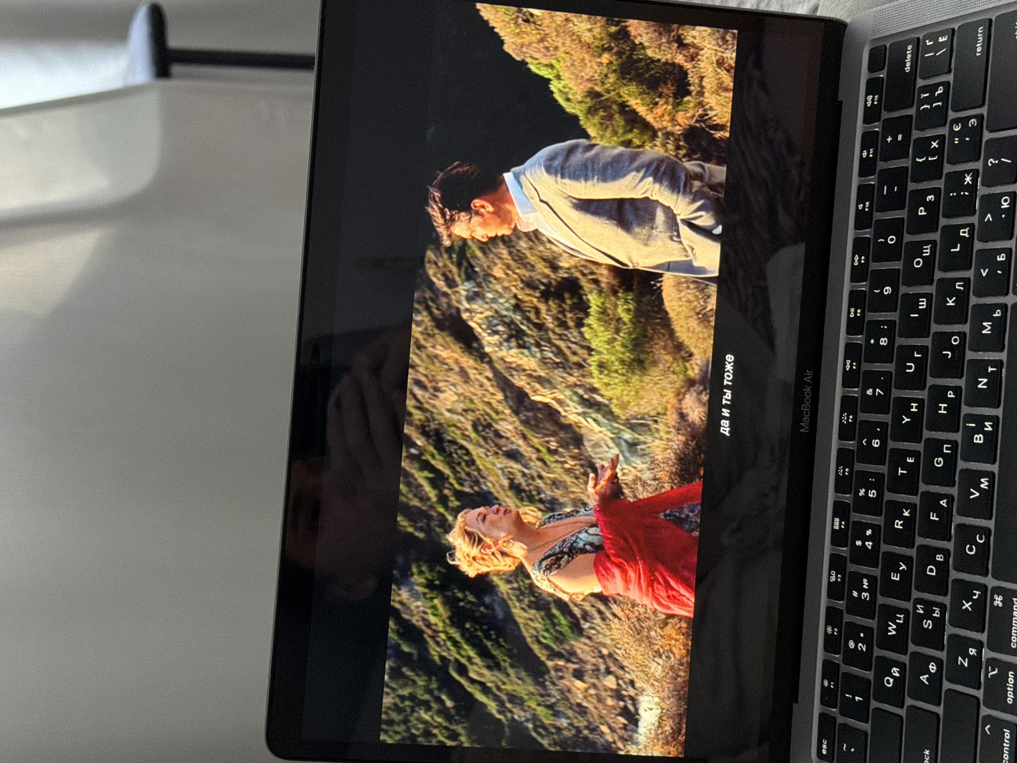 A laptop computer displays a movie scene on its screen. The scene features a blonde woman in a red shawl and a man in a suit standing on a rocky, sunlit hillside covered in dry vegetation. The laptop is open, resting on a flat surface, with its keyboard visible in the foreground. The keyboard is a QWERTY layout, with Cyrillic characters superimposed. The image is taken from a perspective slightly above and to the side of the laptop. The background behind the laptop is blurred, showing a light-colored curtain and a dark chair. There are no visible signs indicating the specific location in Kyiv, Ukraine.