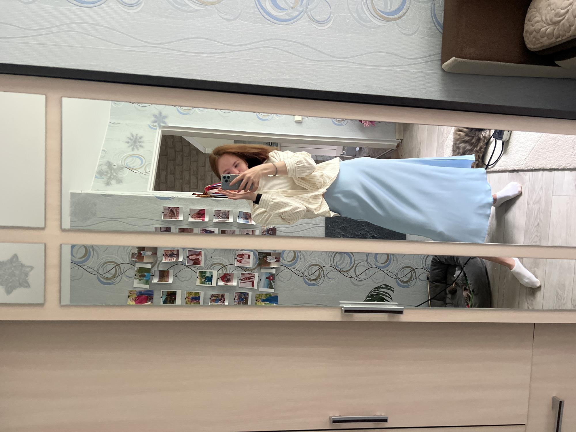 A young woman with auburn hair is taking a mirror selfie in a room. She is wearing a light-colored, striped, long-sleeved top tied at the waist and a light blue, floor-length skirt. She is holding a smartphone in her right hand and looking at her reflection. The mirror reflects a room with patterned wallpaper and a doorway in the background. There are several framed photos attached to the wallpaper to the left of the woman. To the right of the woman, a cat is lying on a rug. The room appears to be a residential interior, likely in an apartment. The woman's pose and the presence of personal photos suggest a casual, personal moment within her home. The overall scene depicts a typical indoor environment. The location is Chaikovsky, Russia, a city known for its industrial facilities, particularly related to energy production, and its residential areas.