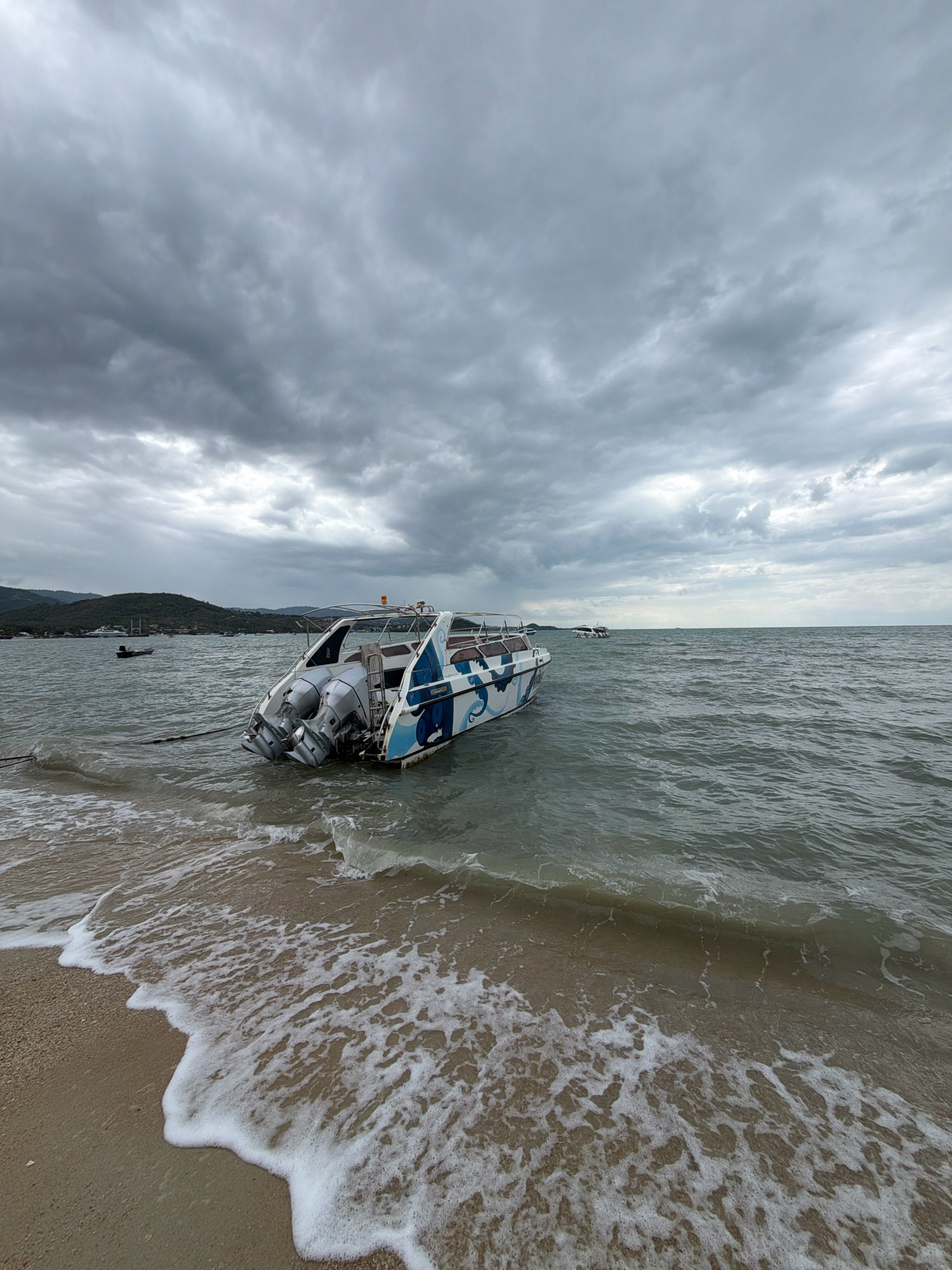 A partially submerged speedboat rests aground on a sandy beach, with its stern facing the viewer and its bow pointing into the choppy, grey sea under an overcast sky. The boat, which is white with blue decorative patterns, has twin outboard motors. The water is disturbed by small waves that lap at the shore. In the background, a distant shoreline with vegetation and a few buildings is visible, along with other boats in the water. The overall scene suggests a cloudy day on the coast of Ko Samui District, Thailand.