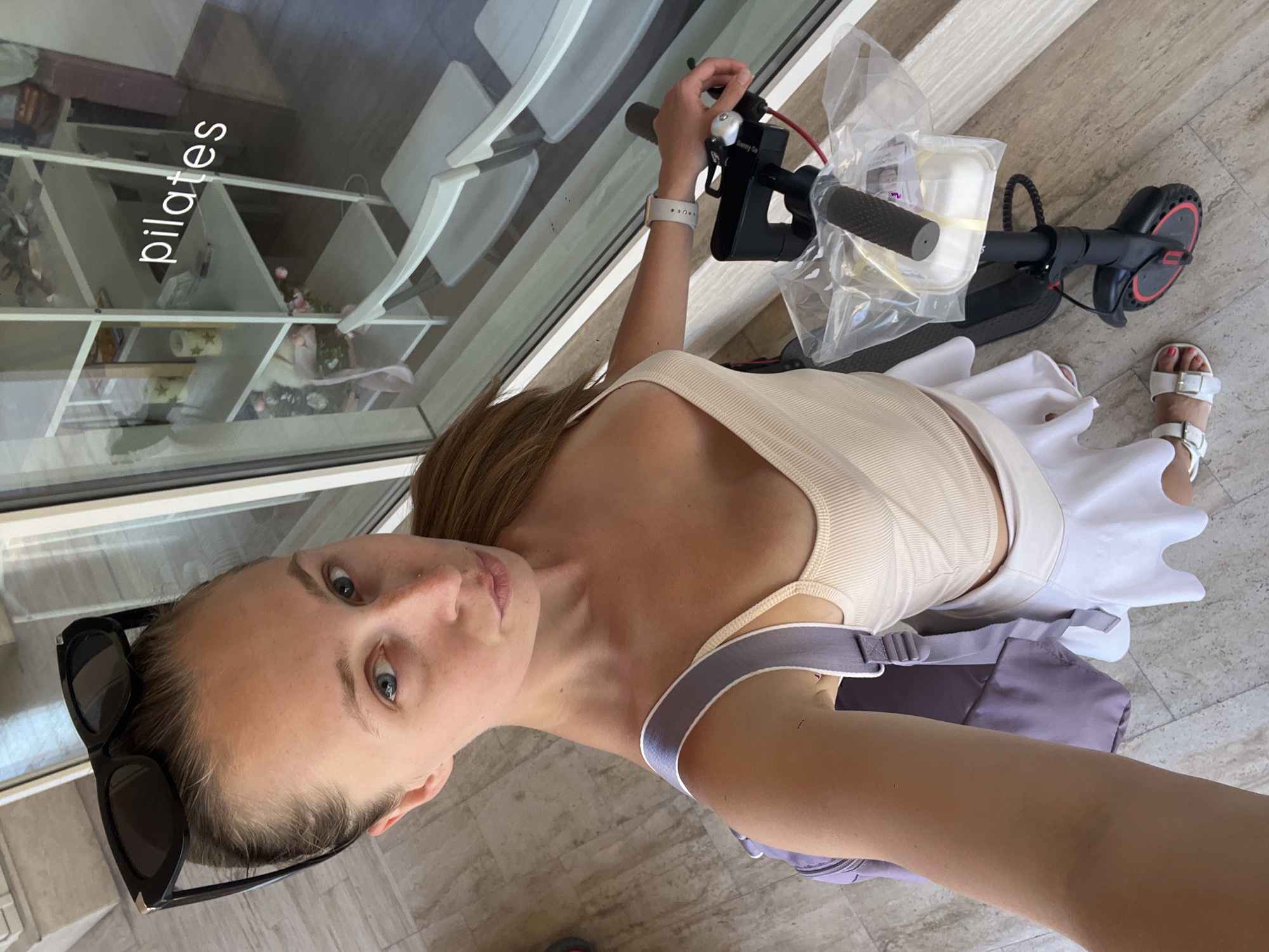 A young woman with blonde hair pulled back and sunglasses perched on her head stands on a tiled floor, facing the camera. She is wearing a beige ribbed tank top and a white pleated skirt. A lavender backpack with grey straps is slung over her left shoulder. To her right, a black electric scooter is parked, with a clear plastic bag containing items secured to the handlebars. The background shows a large window with a reflection of a white shelf unit displaying various items, including candles and decorative flowers. The word "pilates" is visible in white lettering on the glass. The woman appears to be near the entrance of a Pilates studio in Budva, Montenegro.