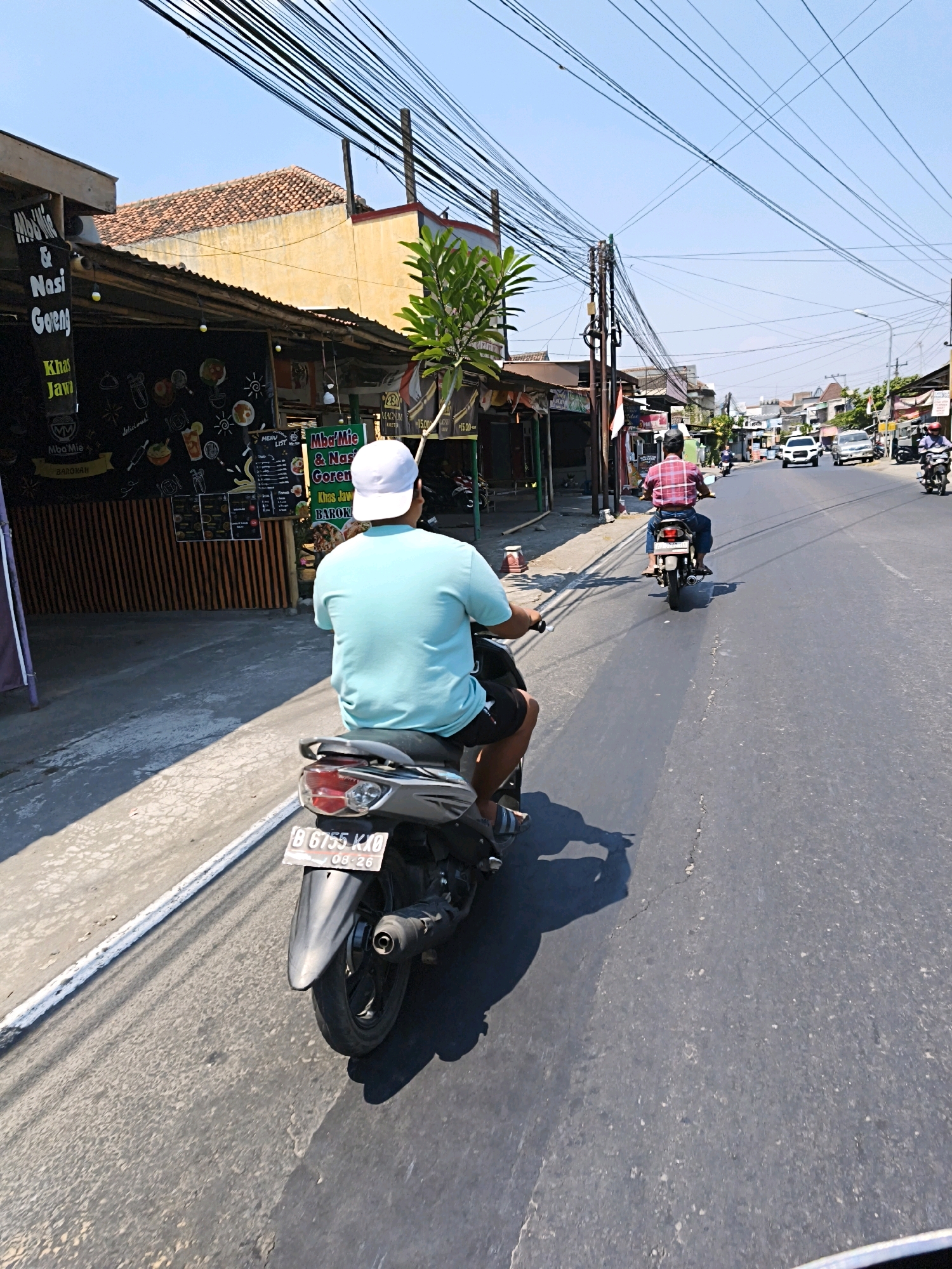 This is a daytime, eye-level shot taken from a moving vehicle, looking down a street in Desa Donohudan, Indonesia. The street is paved and appears to be a local thoroughfare. On the left side of the street, there are several small shops with awnings. One shop has a sign that reads "Mba'Mie & Nasi Goreng". In the foreground, a person is riding a motorcycle. The rider is wearing a light blue t-shirt and dark shorts, and a white baseball cap. The motorcycle has a license plate that reads "B 6755 KXO" and "08-26". Further down the street, another person on a motorcycle is visible, wearing a pink shirt. Several cars and other motorcycles are also on the road. Overhead, a dense network of electrical wires crisscrosses the sky. The overall atmosphere suggests a busy, sunny day in a commercial area of a town or city.
