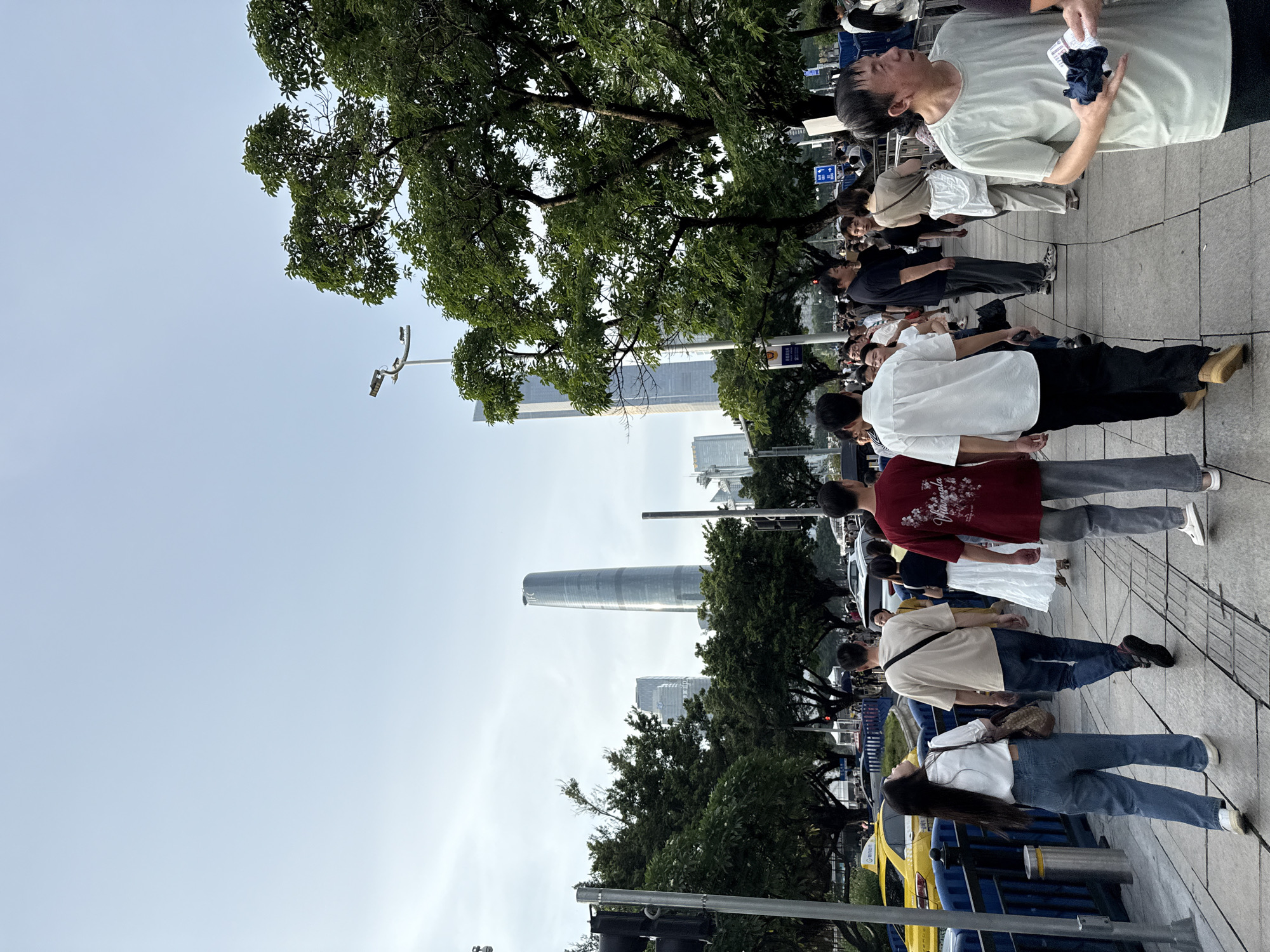 The image depicts a street scene in Haizhu District, China, with several tall, modern buildings visible in the background, including a distinctive, curved skyscraper. A group of people are walking on a sidewalk alongside a street with vehicles, including a yellow taxi. Lush green trees line the sidewalk and street. The sky is overcast. A traffic light is visible on the left side of the frame. Several individuals are captured in motion, walking in various directions, with some appearing to be conversing. The overall atmosphere suggests an urban environment during the daytime.