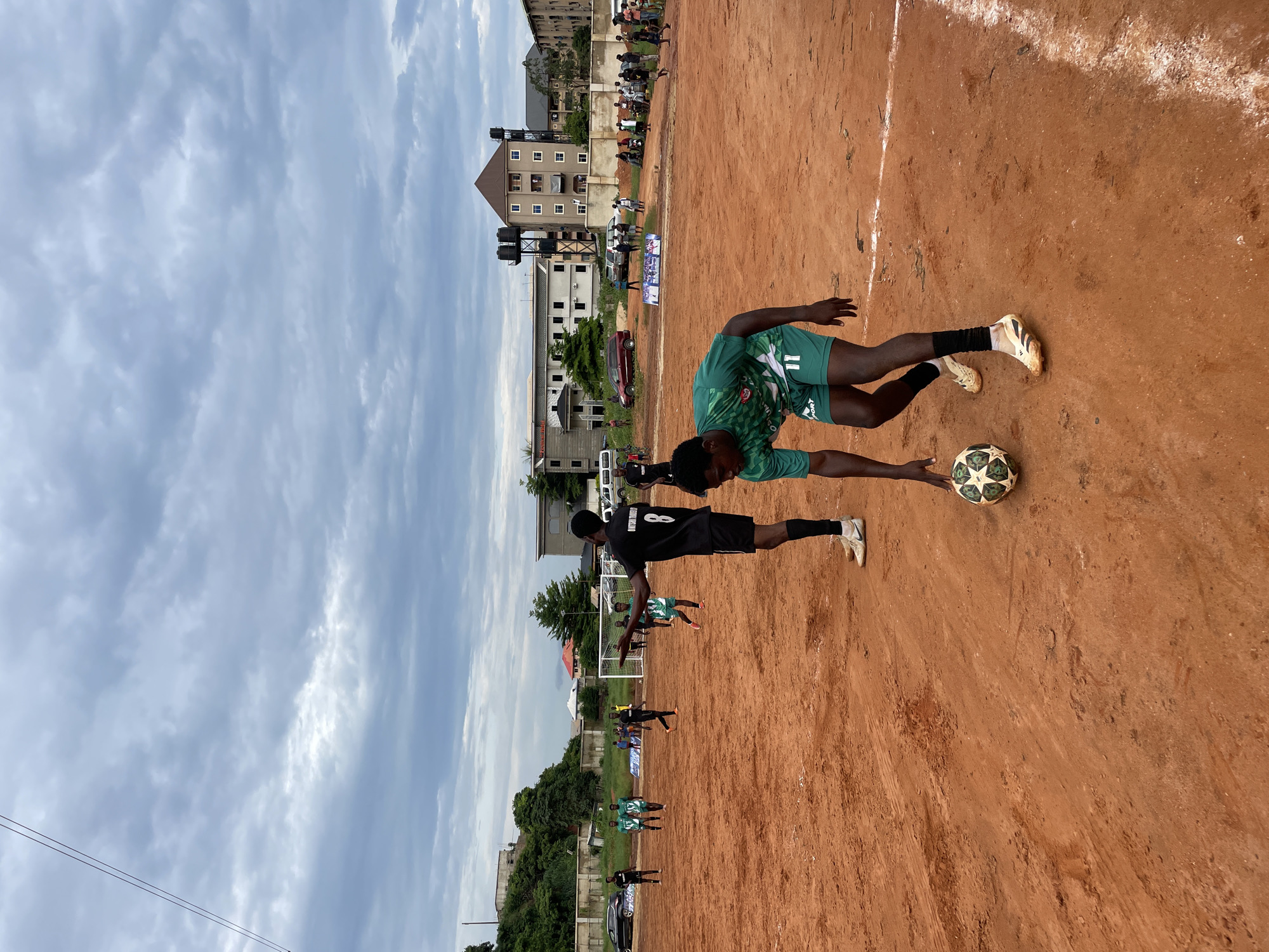 An overcast day features a brown dirt soccer field where a game is in progress. The foreground is dominated by the lower body of a young Black man wearing a green soccer jersey with the number 11 on his shorts, and a white soccer ball near his feet. In the mid-ground, another Black man in a black jersey with the number 8 on his back gestures with his right hand while facing the camera. Other players in green and black jerseys are visible on the field, some running and others standing. A soccer goal is set up at the far end of the field. In the background, buildings of various sizes and styles, some appearing to be multi-story residential or commercial structures, are present. Trees and a fence line the edge of the field, and several cars are parked in the distance. The sky is heavily clouded, with a hazy, diffused light.