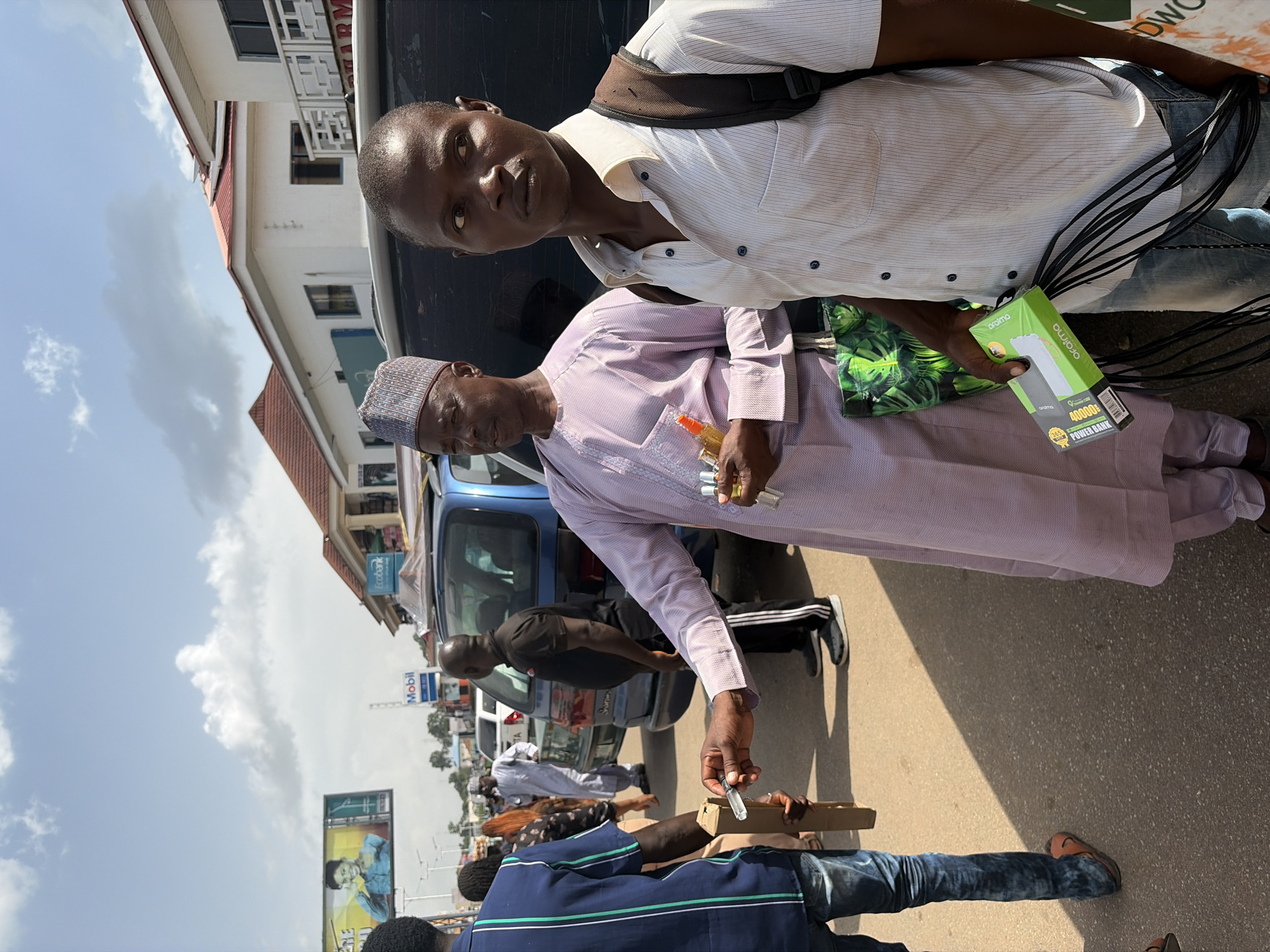 The image shows a street scene in Jos, Nigeria. Two men are the primary subjects. One man, on the left, is wearing a light-colored striped shirt and a dark strap across his chest, possibly a bag strap. He is looking towards the camera with a neutral expression. The second man, in the foreground and slightly to the right, is wearing a pale purple traditional outfit and a patterned cap. He is holding a small object in his right hand. In his left hand, he holds a box that appears to be a power bank, labeled "Oraimo 40000mAh POWER BANK." Several black cables are also visible, connected to the power bank.
Behind them, a blue vehicle is parked. Other people are visible in the background, some walking and others standing. The setting appears to be a roadside market or a busy street. The weather seems to be sunny, with some clouds in the sky, suggesting it is daytime. The overall atmosphere is that of an everyday commercial transaction or interaction on a street. The focus is on the interaction between the two men and the products being displayed or exchanged. Visible text includes "Oraimo" and "40000mAh POWER BANK" on the product box. There are also glimpses of other signs and advertisements in the background, including what appears to be a "Mobil" sign.