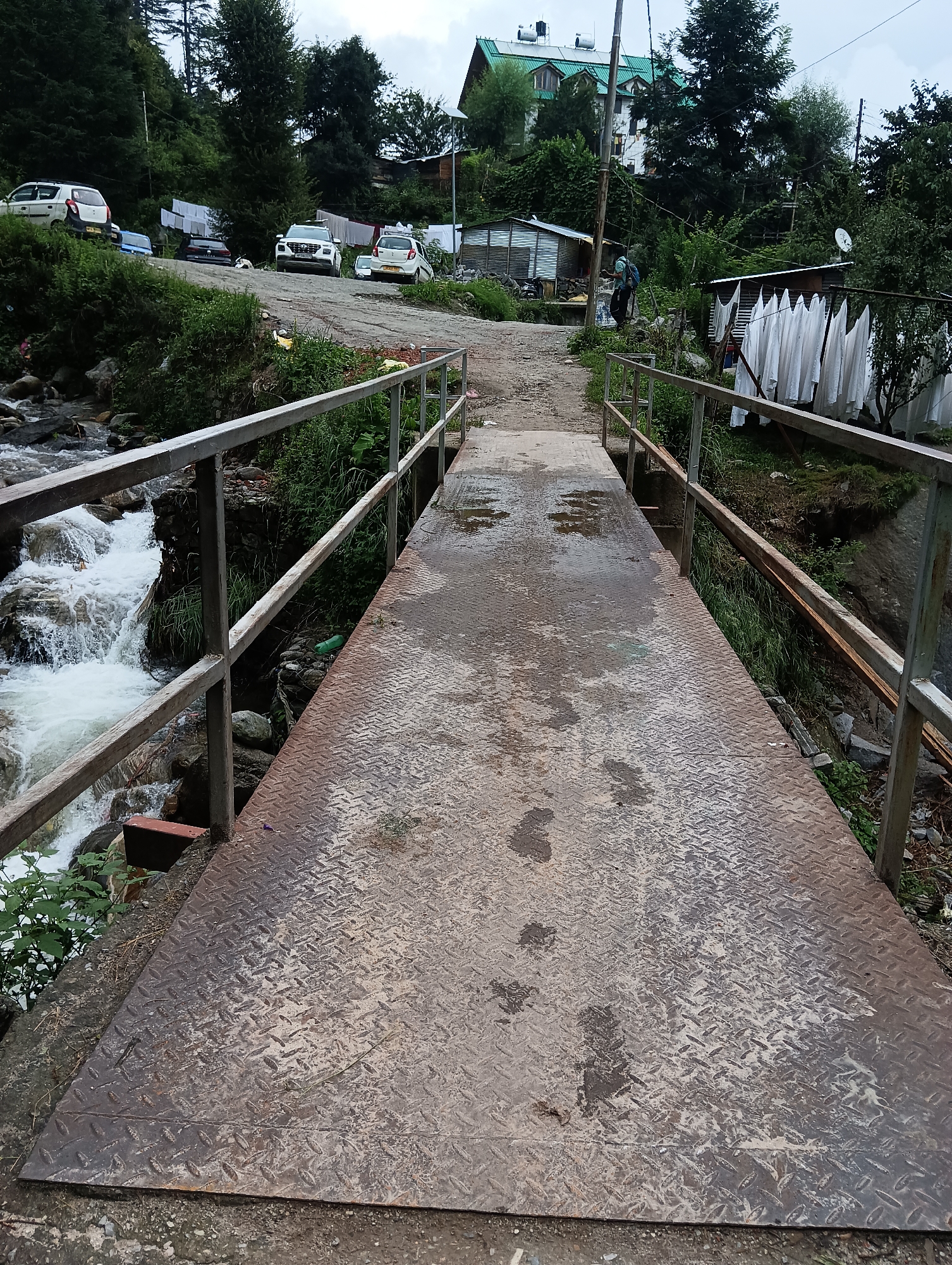 The image depicts a metal pedestrian bridge crossing a fast-flowing river in Siumsa, India. The bridge has diamond plate flooring and metal railings. The riverbanks are overgrown with vegetation. In the background, there is a dirt road leading uphill to a cluster of buildings, including a large house with a green roof and solar panels. Several cars are parked along the road, and laundry is hanging on clotheslines between buildings. A person with a backpack is visible walking uphill on the road. The sky is overcast, suggesting a cloudy day.