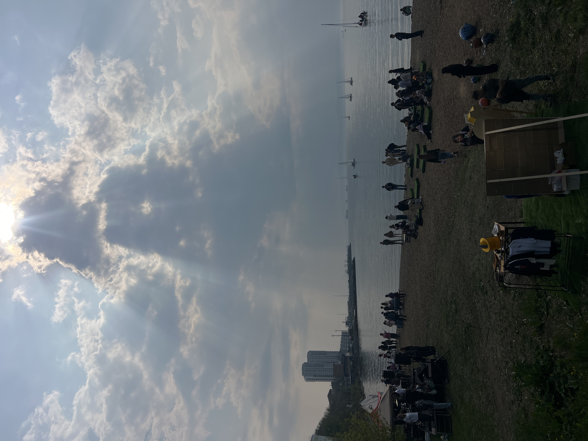 The image depicts a beach scene on what appears to be a partly cloudy day. A large group of people are gathered along the shoreline, some standing near the water, others seated or lying on the beach. Several sailboats are visible in the distance on the water. The image could potentially be taken in Vladivostok, Russia, given the coastal location, the appearance of the buildings, and the climate conditions suggested by the sky. A rack with hanging clothes and a yellow hat is situated on the beach and further in the foreground of the picture, there's a film set in the lower part of the frame.