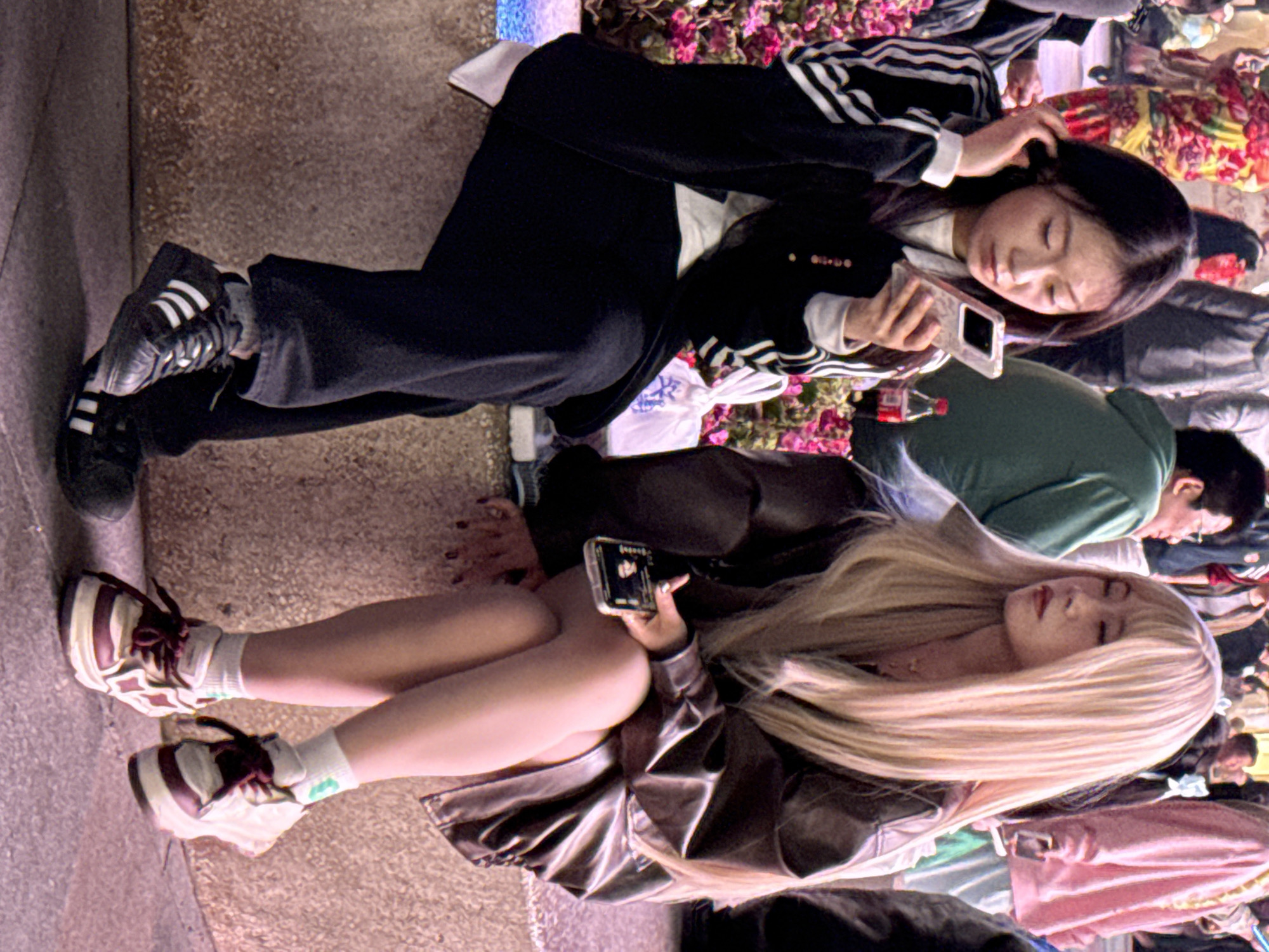 In Liangjiang, China, two young East Asian females are seated on a light-colored, textured concrete ledge in a bustling urban environment. The woman on the left has long, dark hair and is wearing a white long-sleeved shirt under a dark jacket with white stripes on the sleeves, dark pants, and dark Adidas brand sneakers. She holds a silver-colored smartphone in her right hand, gazing at its screen, while her left hand is near her ear. A red soda bottle and a white plastic bag are visible behind her on the ledge, amidst pink and purple flowering plants.
The woman on the right has long, blonde hair and is wearing a dark brown faux leather jacket, exposing her bare legs which are crossed. She wears white socks with a green emblem and white and burgundy/maroon sneakers with black laces. She holds a dark-colored smartphone in her right hand, also looking downwards at its screen, with her eyes appearing closed or downcast. A subtle gold-colored necklace is visible around her neck.
The background is blurred, showing a crowd of diverse individuals, including a person in a green hoodie and another in a pink hoodie, suggesting a public square or pedestrian thoroughfare. The lighting is bright, consistent with an evening or indoor public space. The two women are not directly interacting with each other, both engaged with their personal electronic devices.