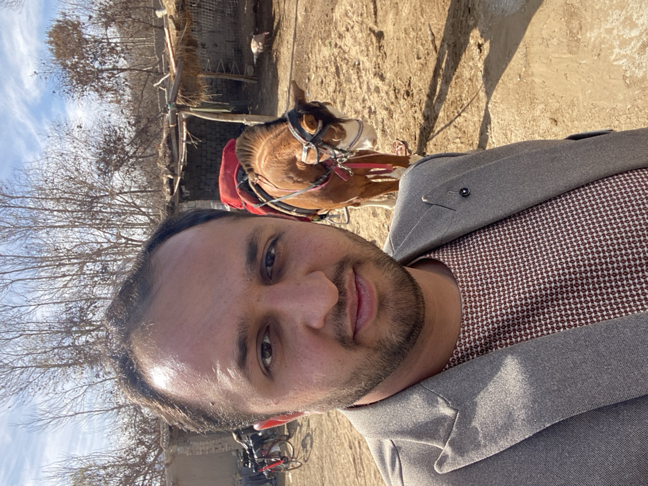 An eye-level, selfie-style photograph captures a male individual in an outdoor, semi-rural setting in Kandahar, Afghanistan. The subject, a man with light-brown skin, short dark hair, and a short beard, occupies the left half of the frame, looking directly at the camera with a neutral expression. He wears a light grey blazer over a dark red/brown shirt with a small geometric pattern; a black pin is affixed to his left lapel. Behind him, slightly to the right, a brown horse stands still, fitted with a red padded saddle and a bridle with white and black detailing. In the far left background, two bicycles are visible, one red, leaning against a low, light-colored wall. Further back, a dilapidated mud-brick structure and a rustic shelter with a straw roof supported by wooden poles are present. A light-colored bird, possibly a turkey or chicken, is visible on the unpaved, dusty light-brown ground near the shelter. The background consists of numerous deciduous trees with bare branches under a partly cloudy blue sky. Sunlight casts distinct shadows on the ground.