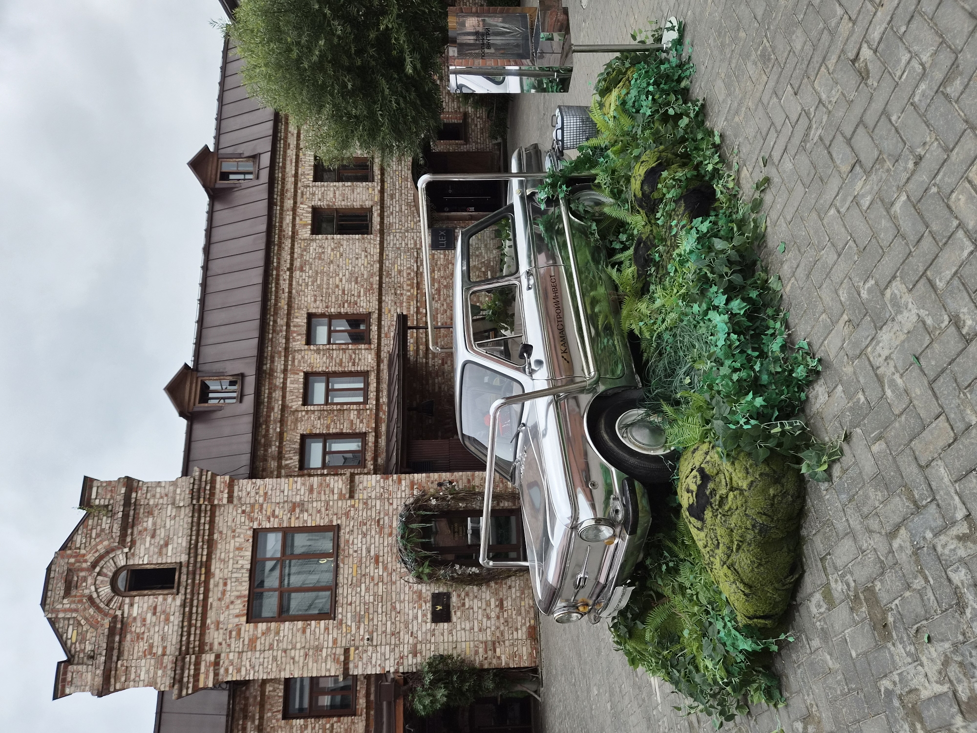 The image depicts an outdoor courtyard scene at the Kazan Clock Museum, located in Kazan, Russia. The central focus is a vintage, chrome-plated car, possibly a Zaporozhets, displayed amidst an artificial landscape of lush green foliage and large moss-covered rocks. The car is positioned on a cobbled pavement.
Behind the car, a historic brick building with a dark, sloped roof stands prominently. The building features multiple windows with dark frames, some of which are open. A sign with the text "ZUCKERS" is visible near an arched entrance adorned with greenery. Further to the right, another section of the brick building is visible, along with a mature tree.
A reflective panel, resembling a mirror, is installed on a pole to the right of the car. It reflects the surrounding environment and part of the car's interior. A cylindrical trash bin is also present near the car. The sky is overcast, suggesting a cloudy day. No people are visible in the scene.