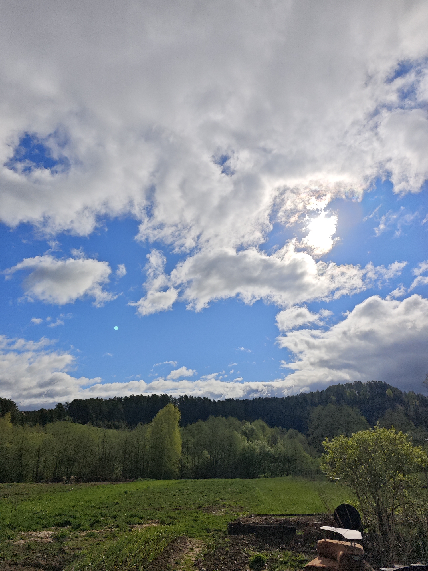 The image presents a rural scene in Фоки, Russia, characterized by a bright, partly cloudy sky with the sun visible through a layer of clouds. Below the sky, a densely forested hill dominates the mid-ground, its trees varying in shades of green. In front of the forest, a grassy field extends towards the viewer, with patches of bare earth visible in the foreground. A shrub with green leaves is situated on the right side of the frame. The bottom-right of the frame shows some bricks with a metallic plate on them.