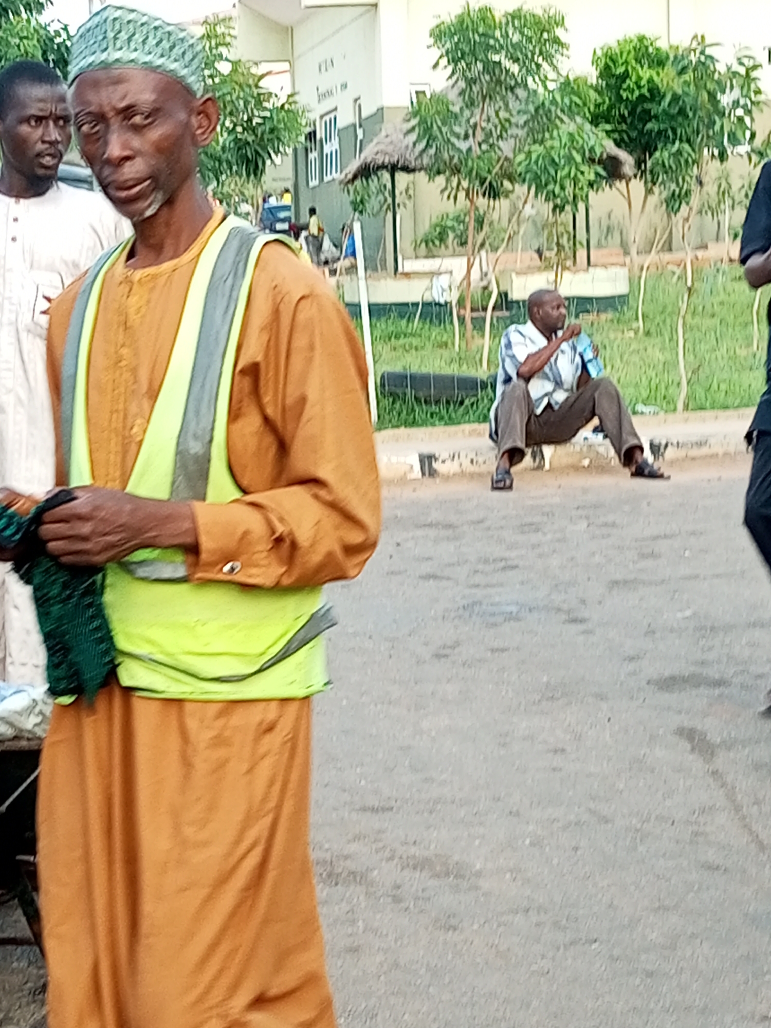 In the foreground, a man wearing a light green cap, an orange robe, and a reflective yellow vest stands looking to his left, holding a green cloth in his right hand. Behind him, a second man in a white robe with a dark pattern on the chest stands looking forward with his mouth slightly open. In the mid-ground, a third man sits on a curb wearing a blue and white striped shirt, brown pants, and dark sandals, holding a light blue object. He is looking to his right. The background shows a building with windows, trees, and grass. The ground is paved with asphalt. The scene appears to be an outdoor public space.