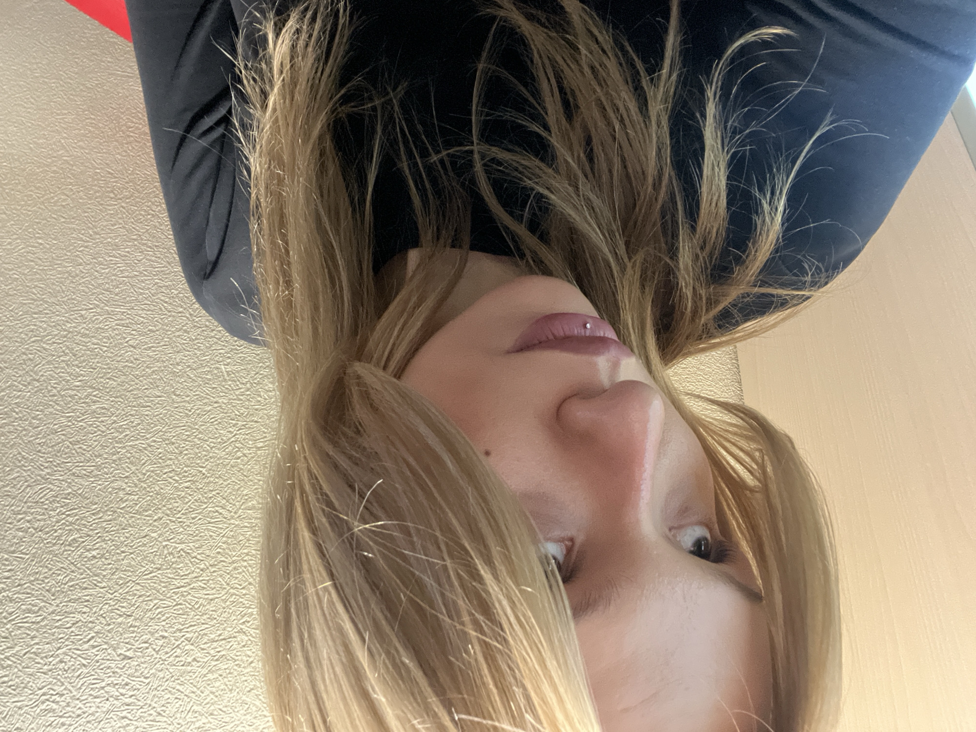 A close-up, upside-down selfie shows a woman with light brown hair and fair skin. She wears a black, long-sleeved shirt. Her lips are slightly parted and appear to have a piercing. One eye is partially visible. The background includes textured, light-colored walls. The location of Chaikovsky, Russia, cannot be determined from this image.